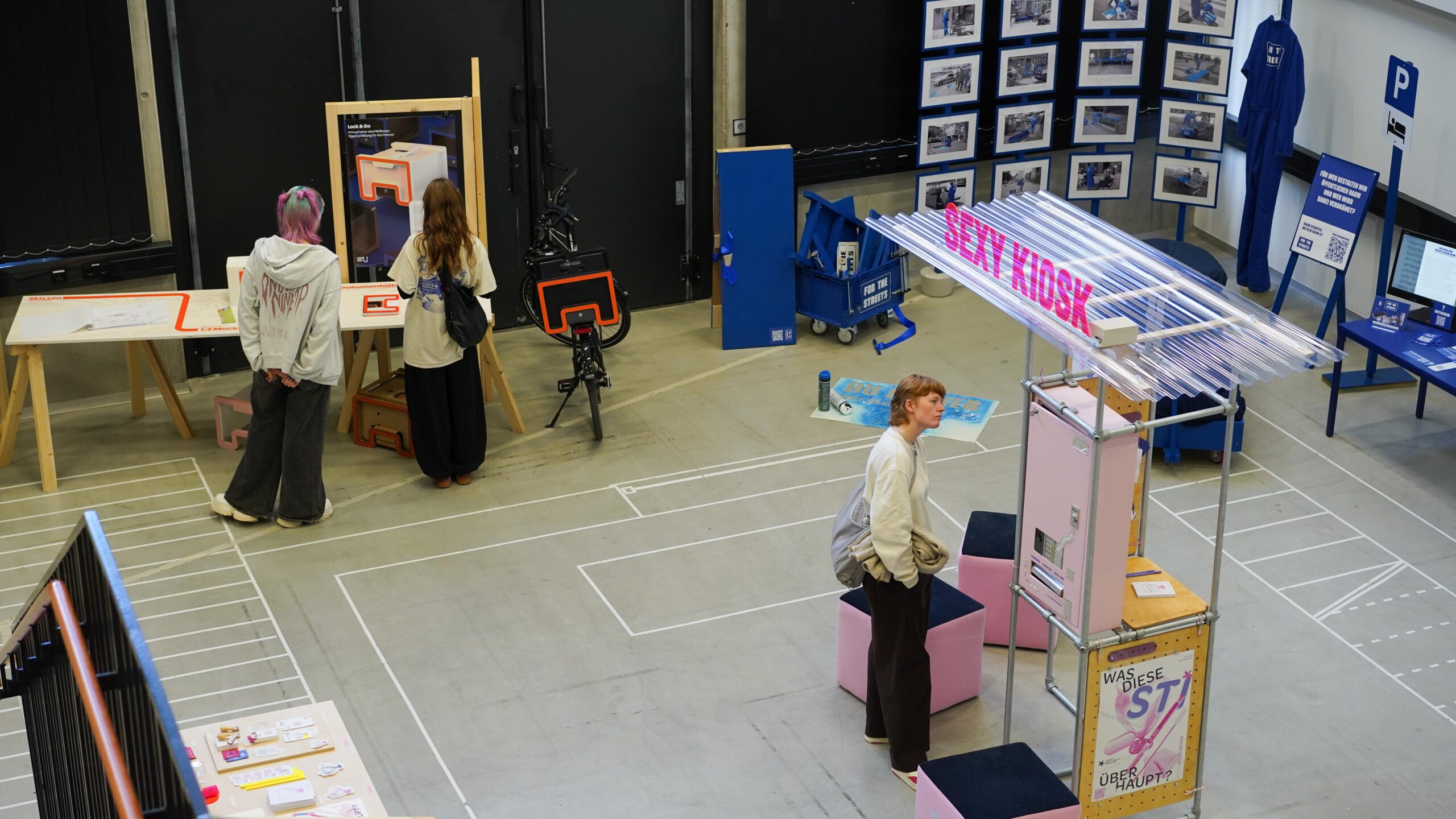 Drei Personen betrachten Exponate in einem modernen Ausstellungsraum, der mit Informationsplakaten, Kunstwerken und interaktiven Installationen ausgestattet ist. Ganz im Sinne einer Werkschau umfasst die Szene sorgfältig angelegte Wege auf dem Boden sowie farbenfrohe Ausstellungsstände, die innovatives Design und kreative Ansätze im Bereich „Bauen“ präsentieren. at Hochschule Coburg