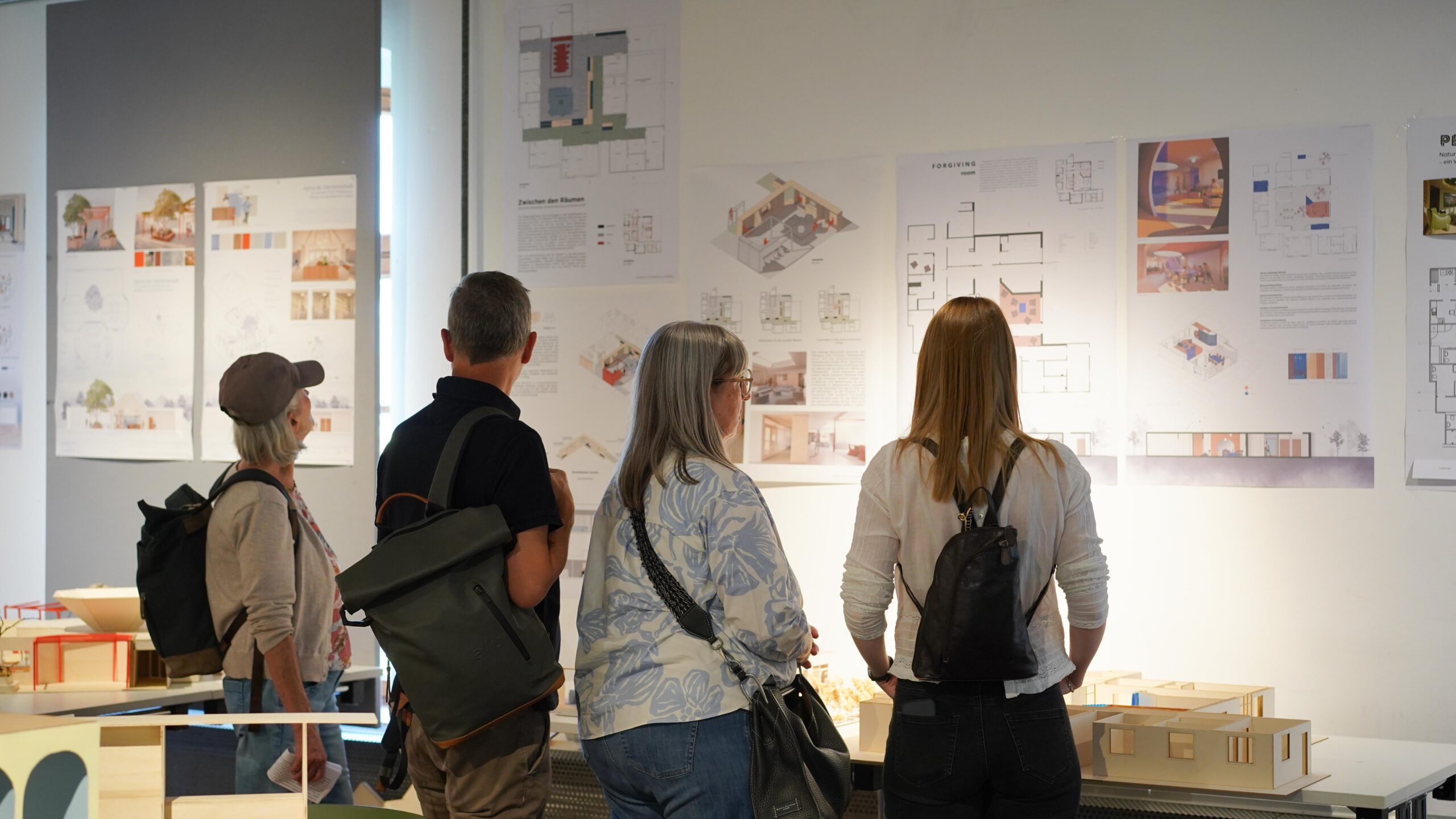 Vier Personen mit Rucksäcken stehen dicht beieinander und betrachten Architekturzeichnungen und Gebäudemodelle, die an einer Wand in einem hell beleuchteten Ausstellungsraum ausgestellt sind – ein Ausdruck des innovativen Geistes des Designs auf der Werkschau für Bauen. at Hochschule Coburg