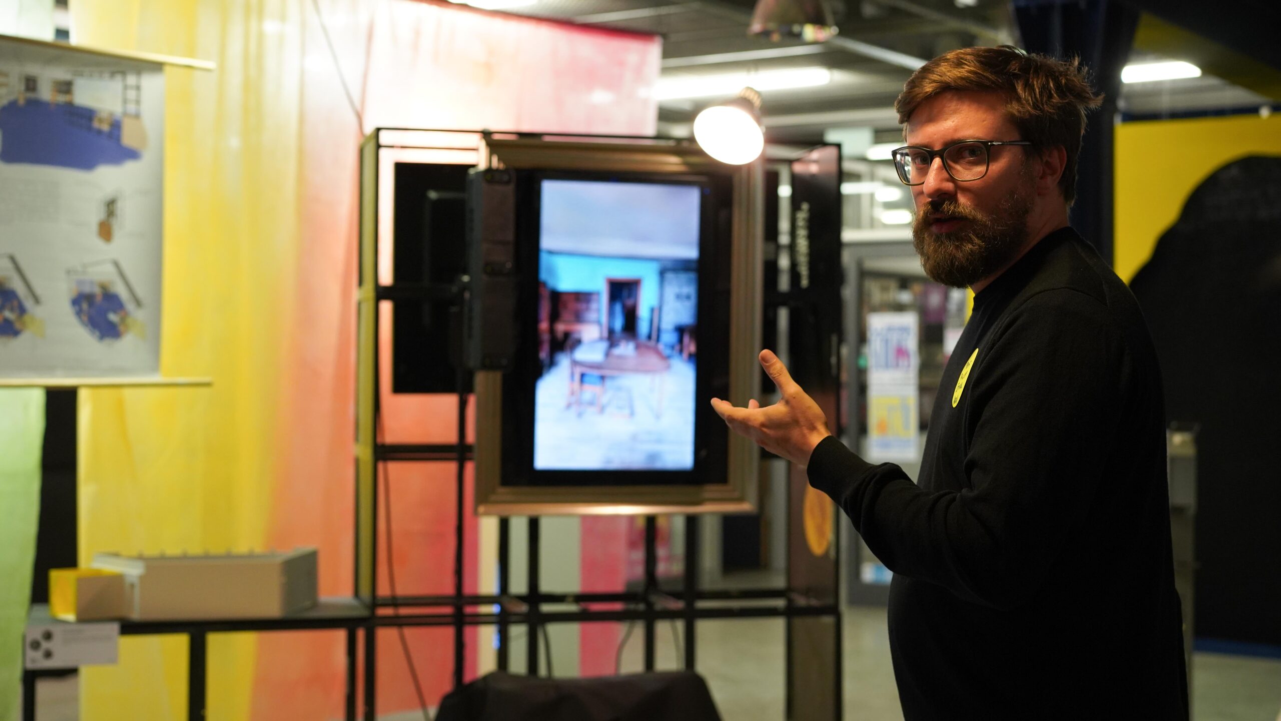 Ein bärtiger Mann mit Brille deutet auf einen digitalen Bildschirm, auf dem das Interieur eines Raums zu sehen ist. Er steht in einem Ausstellungsraum mit bunten Vorhängen und Diagrammen an den Wänden – eine Szene, die die innovative Energie des zeitgenössischen Designs auf dieser Werkschau einfängt. at Hochschule Coburg