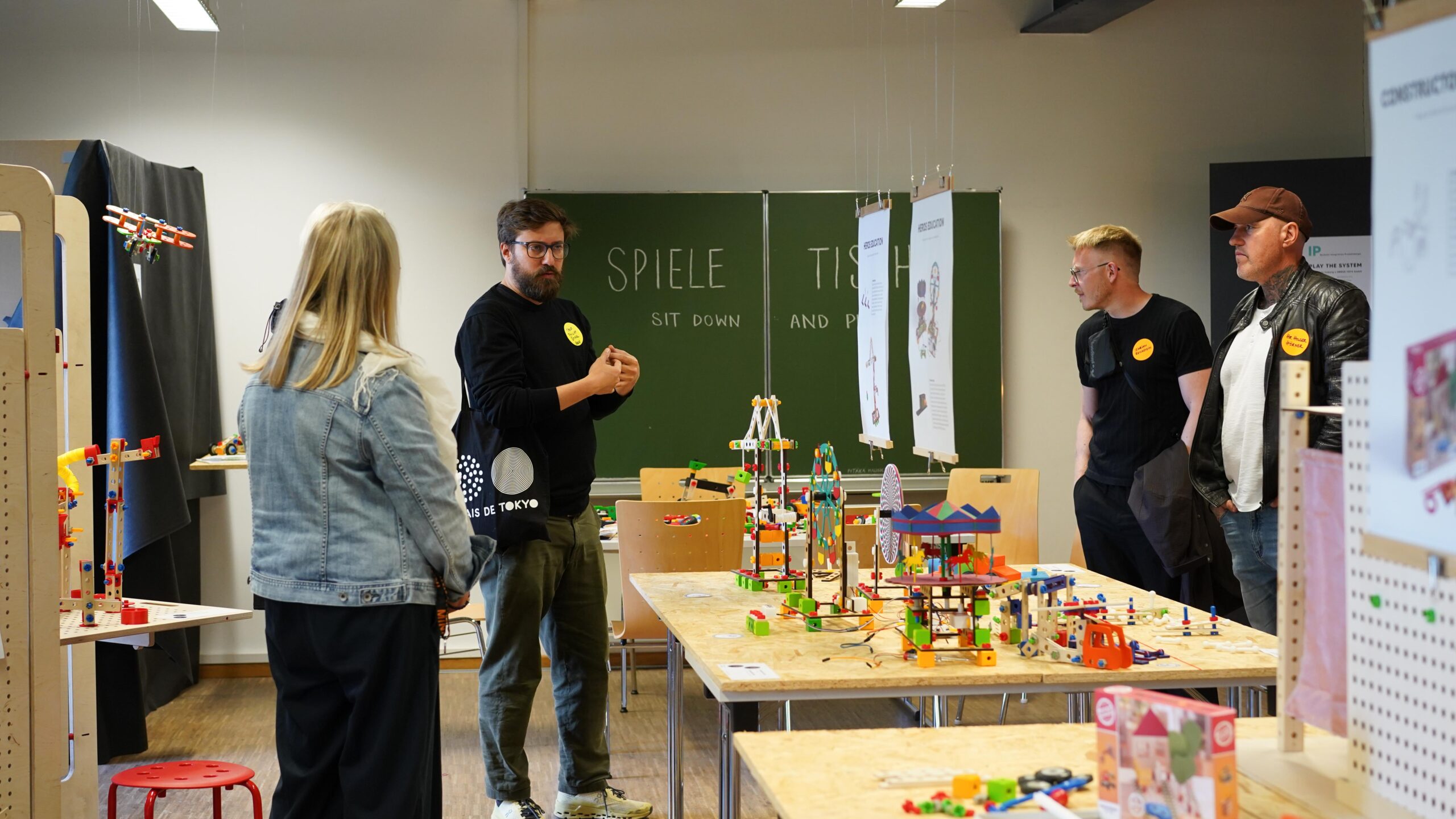 Vier Erwachsene stehen in einem klassenzimmerähnlichen Raum um Tische herum, auf denen buntes Bauspielzeug ausgestellt ist, und führen eine lebhafte Werkschau zum Thema kreatives Design und Bauen durch. Ein Mann spricht, während die anderen aufmerksam zuhören. Im Hintergrund ist eine grüne Tafel mit Schriftzügen zu sehen. at Hochschule Coburg
