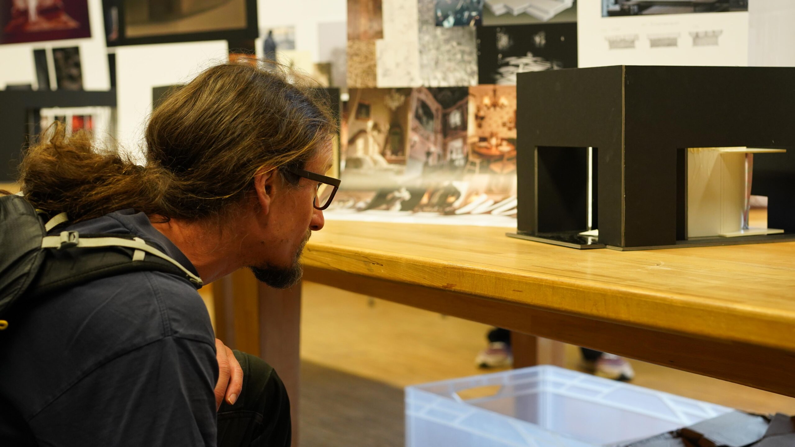 Eine Person mit langen Haaren und Brille betrachtet aufmerksam ein schwarzes Architekturmodell auf einem Holztisch und stellt dabei inmitten der kreativen Atmosphäre einer Werkschau – vor dem Hintergrund von Entwurfsplänen und Bildern – ihr gestalterisches Können unter Beweis. at Hochschule Coburg