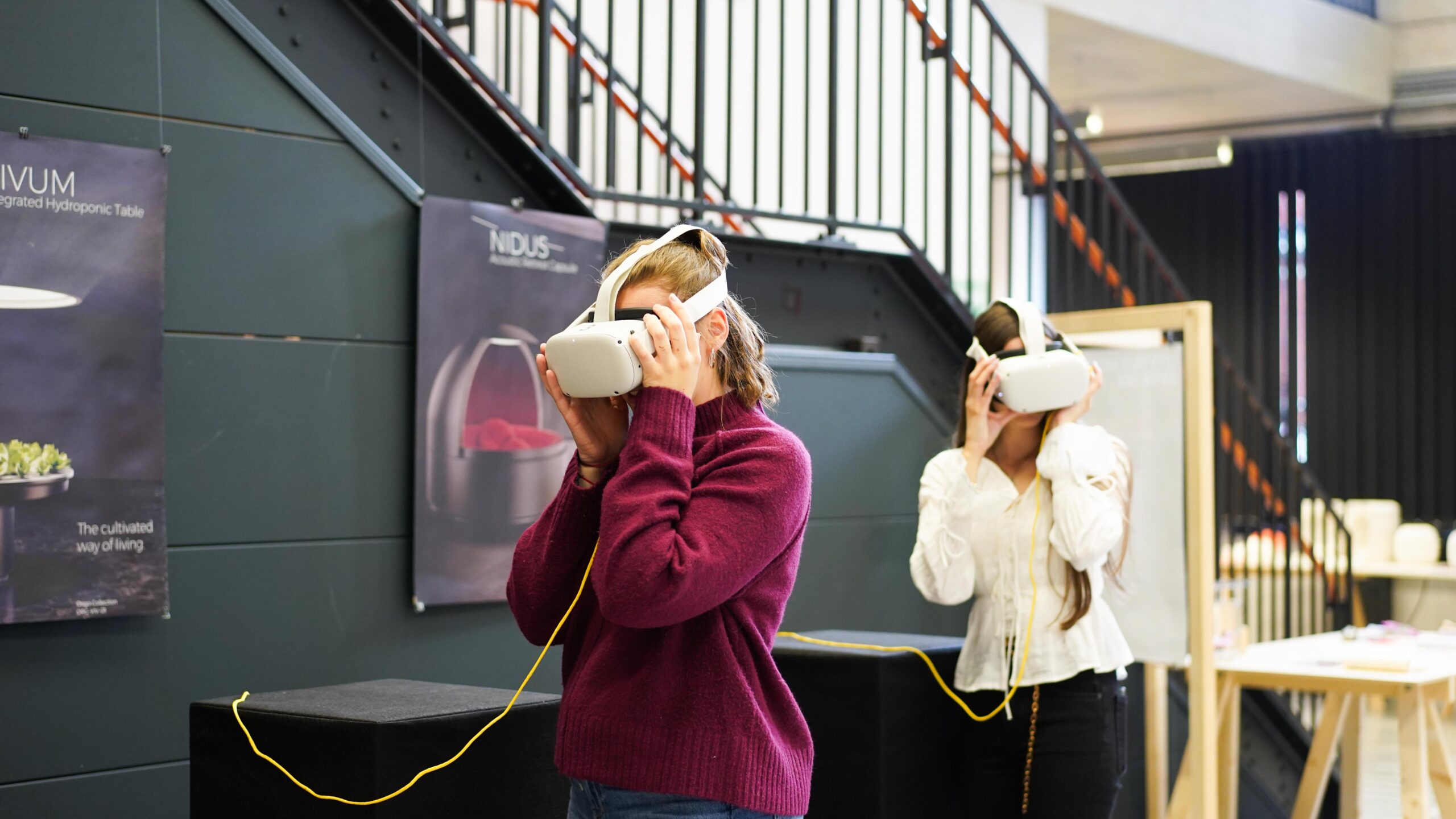 Zwei Personen mit VR-Headsets stehen in einem Innenraum und sind ganz in ein virtuelles Designerlebnis vertieft. Die Person im Vordergrund trägt einen kastanienbraunen Pullover, die Person im Hintergrund einen weißen. Beide halten die Headsets mit den Händen fest. Im Hintergrund sind eine Treppe und Plakate zu sehen – vielleicht Hinweise auf eine kürzlich stattgefundene Werkschau. at Hochschule Coburg