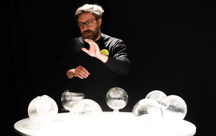 Ein Mann mit Brille deutet auf einen runden weißen Tisch, auf dem mehrere durchsichtige Glaskugeln ausgestellt sind, die als Teil einer Werkschau ein einzigartiges Design präsentieren und vor einem dunklen Hintergrund stehen. at Hochschule Coburg