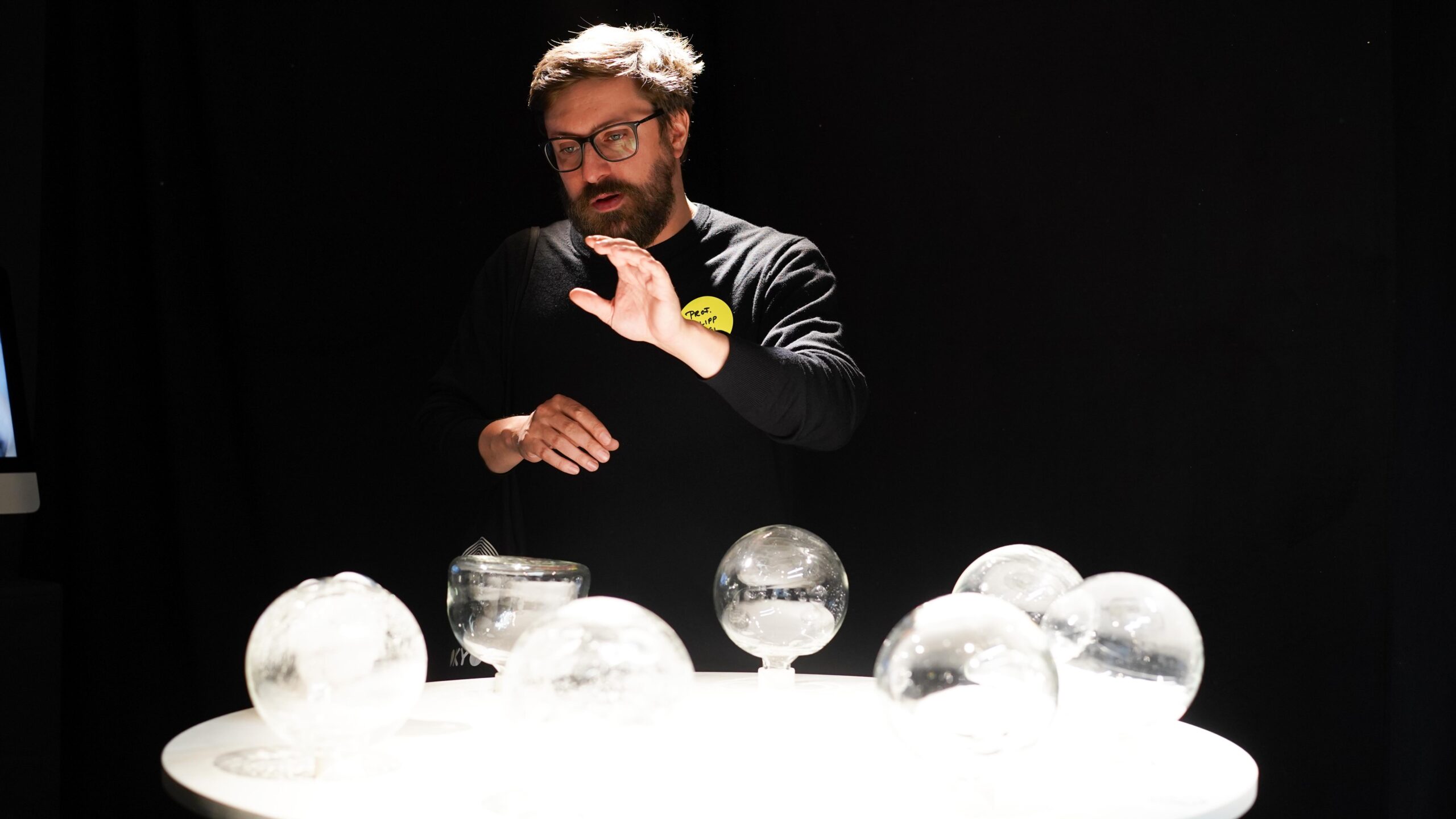 Ein Mann mit Brille deutet auf einen runden weißen Tisch, auf dem mehrere durchsichtige Glaskugeln ausgestellt sind, die als Teil einer Werkschau ein einzigartiges Design präsentieren und vor einem dunklen Hintergrund stehen. at Hochschule Coburg