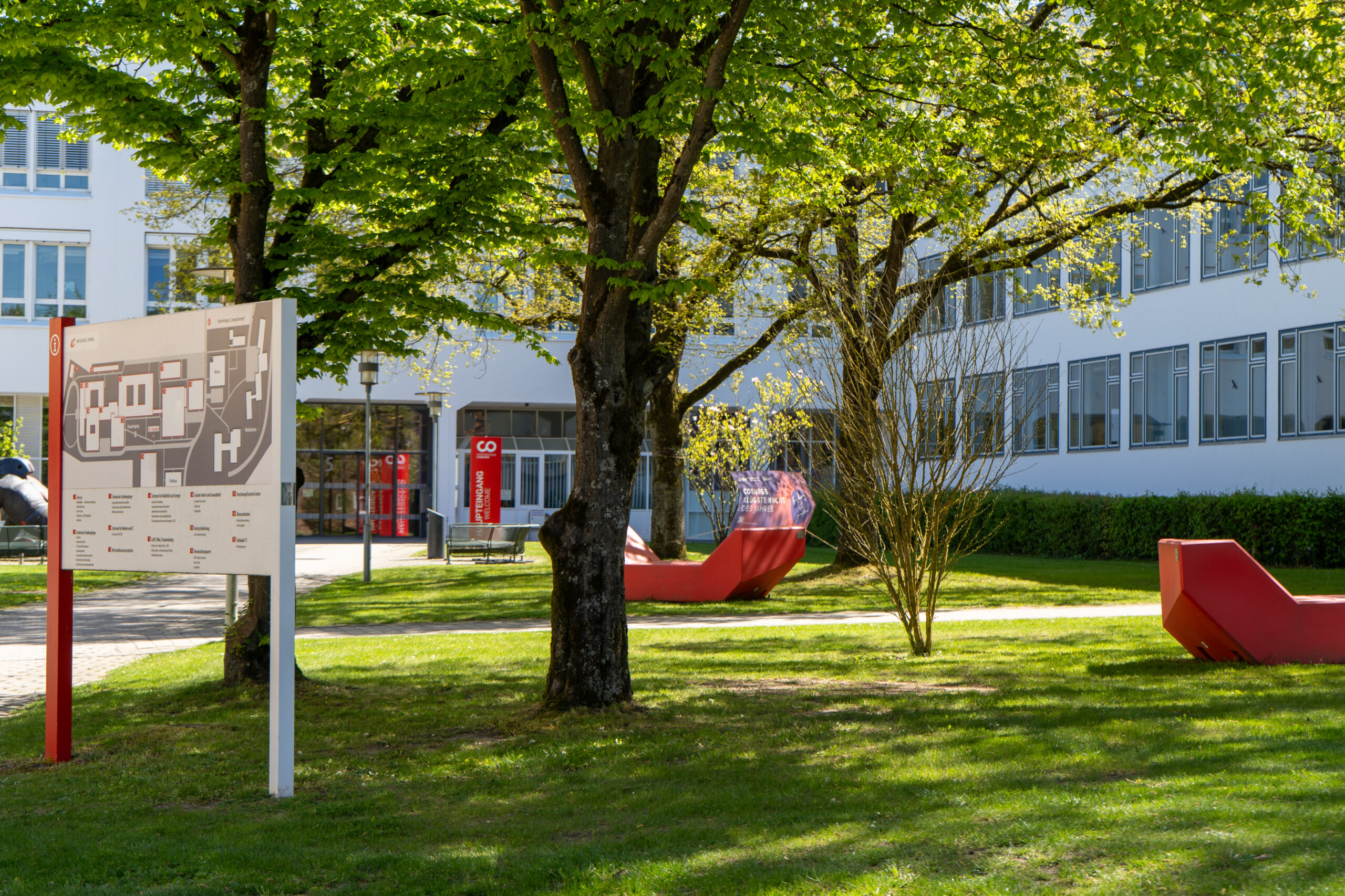 Ein sonniger Innenhof mit grünem Rasen, Bäumen, einem Schild mit dem Campusplan, roten Bänken und einem modernen Gebäude mit vielen Fenstern im Hintergrund bietet eine inspirierende Kulisse für Studierende, die ihren Karriereweg im Rahmen der Professur erkunden. at Hochschule Coburg