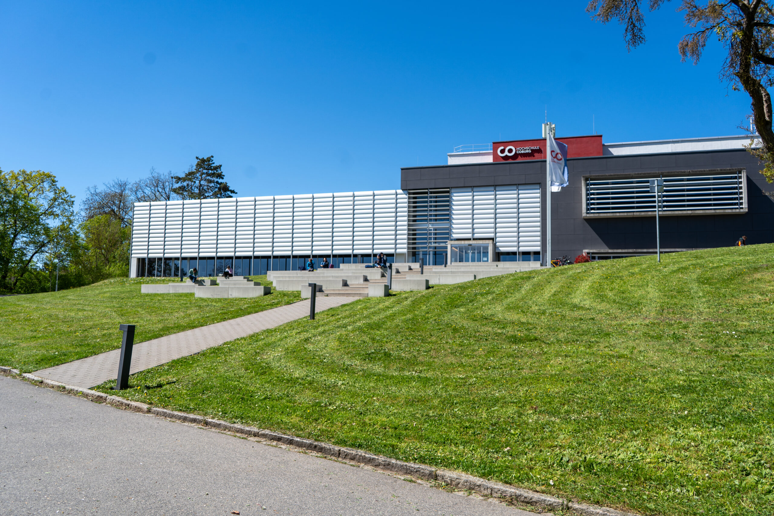 Ein modernes Gebäude mit großen Fenstern und einem roten Akzent steht auf einem grasbewachsenen Hügel unter strahlend blauem Himmel. Stufen und ein Weg führen zum Eingang und strahlen eine einladende Atmosphäre für diejenigen aus, die ihren Karriereweg einschlagen möchten. In der Nähe steht ein Fahnenmast, der neue Möglichkeiten für akademisches Wachstum oder eine Professur innerhalb dieser Mauern symbolisiert. at Hochschule Coburg