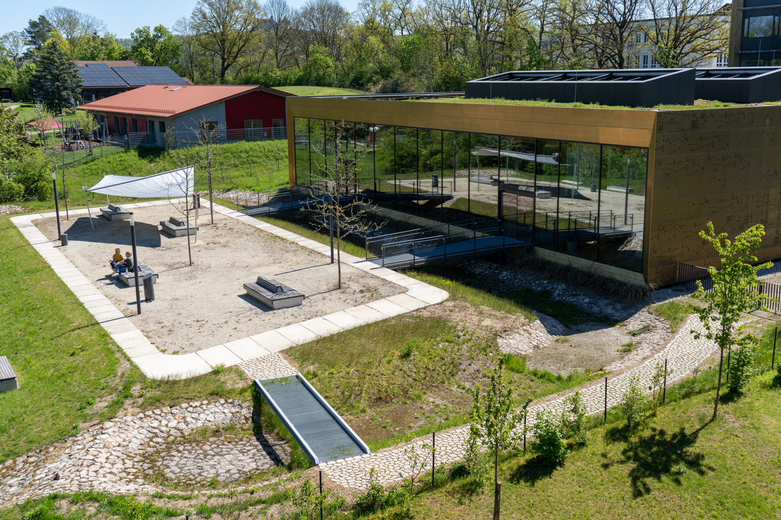 Ein modernes Gebäude mit großen Glasfenstern – ideal für alle, die eine Karriere als Professor anstreben – steht neben einem sandigen Spielplatz mit Bänken und einem Sonnendach. In der Nähe überquert eine kleine Metallbrücke einen felsigen Bach, der von Gras und jungen Bäumen umgeben ist, und schafft so eine inspirierende Kulisse für akademisches Wachstum. at Hochschule Coburg