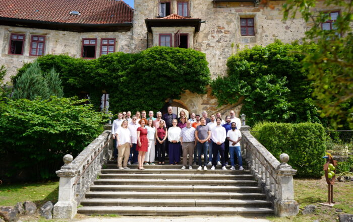 Eine Gruppe von Menschen posiert für ein Foto auf einer Steintreppe vor einem historischen, von Weinreben umrankten Gebäude mit roten Fenstern und üppigem Grün – ganz im Sinne des Austauschs und zur Würdigung guter Doktorarbeiten im Rahmen der Promotionstagung. at Hochschule Coburg