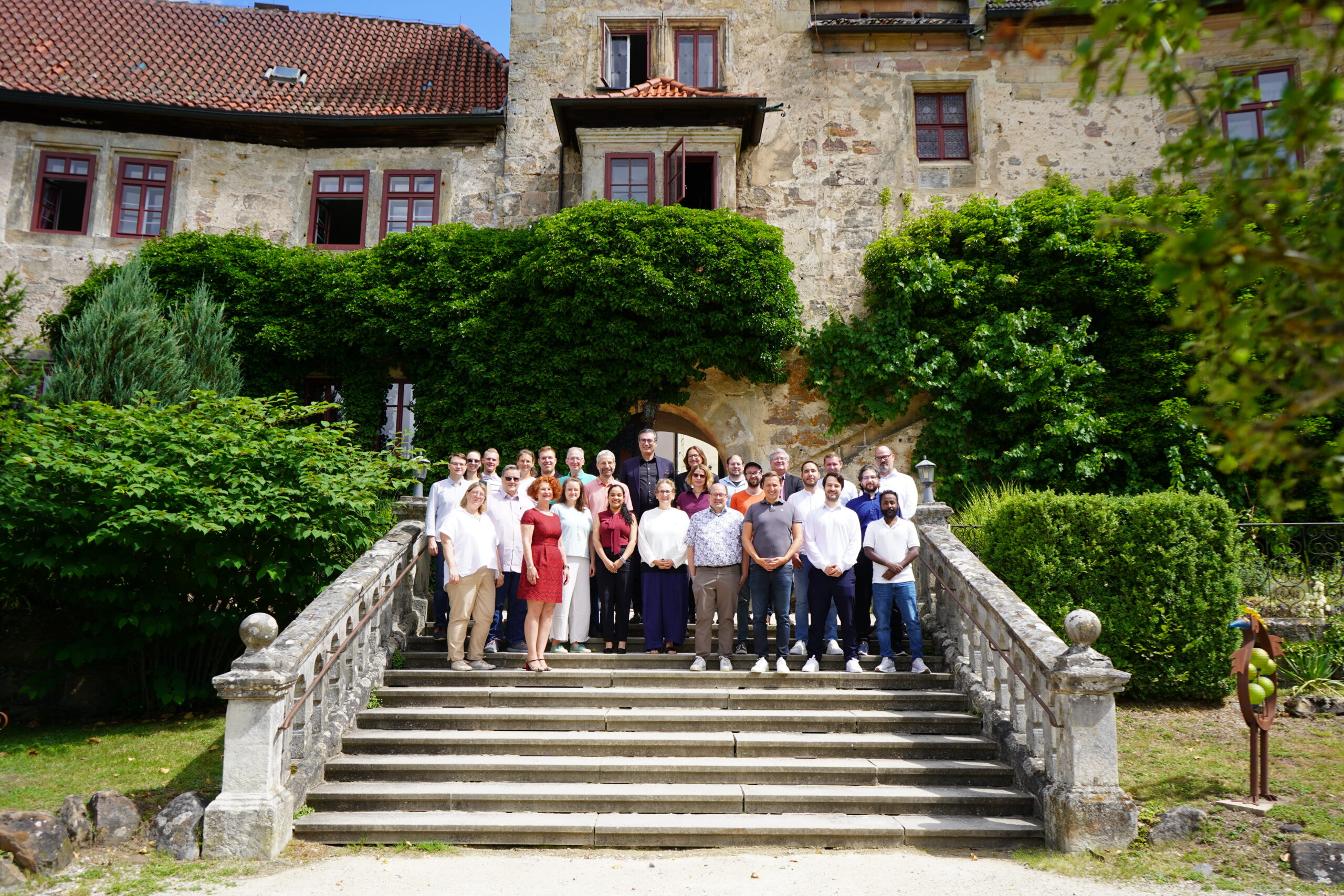 Eine Gruppe von Menschen posiert für ein Foto auf einer Steintreppe vor einem historischen, von Weinreben umrankten Gebäude mit roten Fenstern und üppigem Grün – ganz im Sinne des Austauschs und zur Würdigung guter Doktorarbeiten im Rahmen der Promotionstagung. at Hochschule Coburg