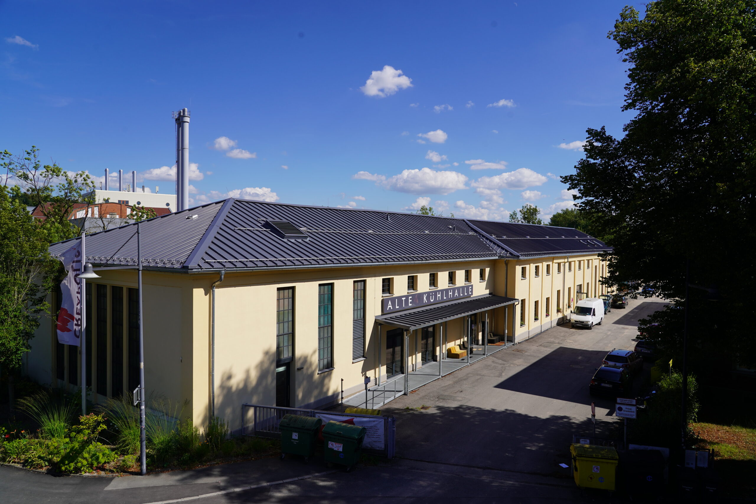 Ein langes, hellgelbes Gebäude mit grauem Dach und der Aufschrift „Alte Kühlhaus“ steht unter einem blauen Himmel mit vereinzelten Wolken, umgeben von Bäumen und geparkten Fahrzeugen – eine passende Kulisse für akademische Veranstaltungen, Diskussionen über die akademische Karriere oder für diejenigen, die eine Professur anstreben. at Hochschule Coburg