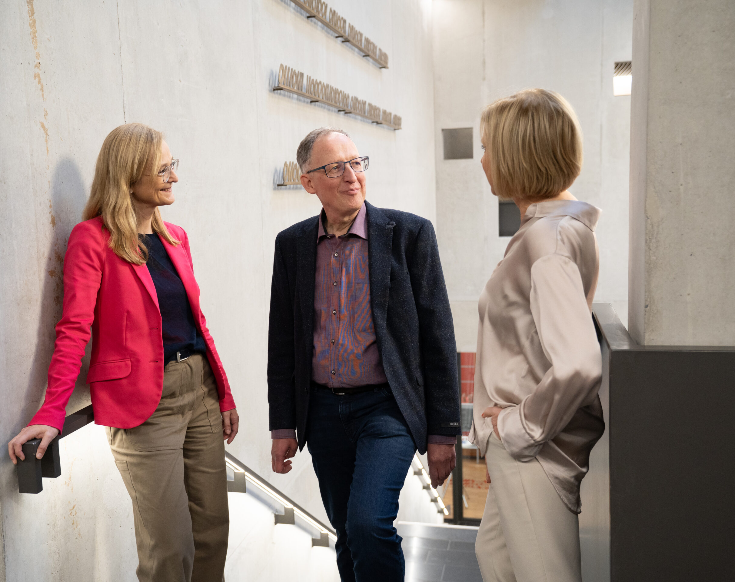Drei Erwachsene stehen auf einer modernen Innentreppe und unterhalten sich, vielleicht über ihren Karriereweg. Zwei Frauen – eine in einem rosa Blazer, die andere in einer beigen Bluse – stehen einem Mann mit Brille und blauem Blazer gegenüber. Die helle Kulisse mit Betonwänden vermittelt den Eindruck einer inspirierenden Umgebung – perfekt für Gespräche über Karrierewege oder das Streben nach einer Professur. at Hochschule Coburg