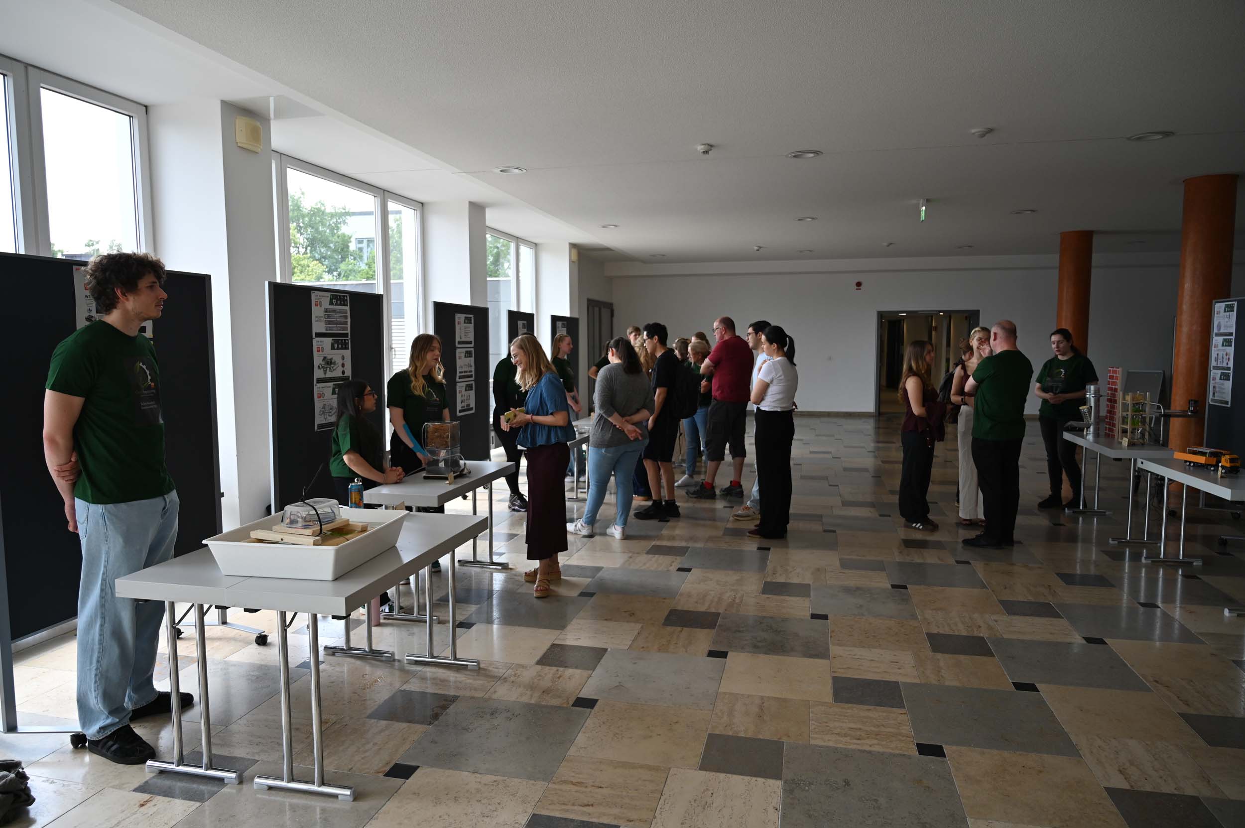 Die Menschen versammeln sich in einem hellen Saal, in dem auf Tischen Modelle und Plakate zum Thema „Lehre“ und zur Bedeutung von „Wasser retten“ ausgestellt sind. Einige stehen hinter den Tischen und tauschen sich mit anderen über ihre Studienerfahrungen aus, während die Besucher die Exponate betrachten und sich in kleinen Gruppen unterhalten. Eine Wand ist mit großen Fenstern versehen, die den Raum mit Tageslicht durchfluten. at Hochschule Coburg