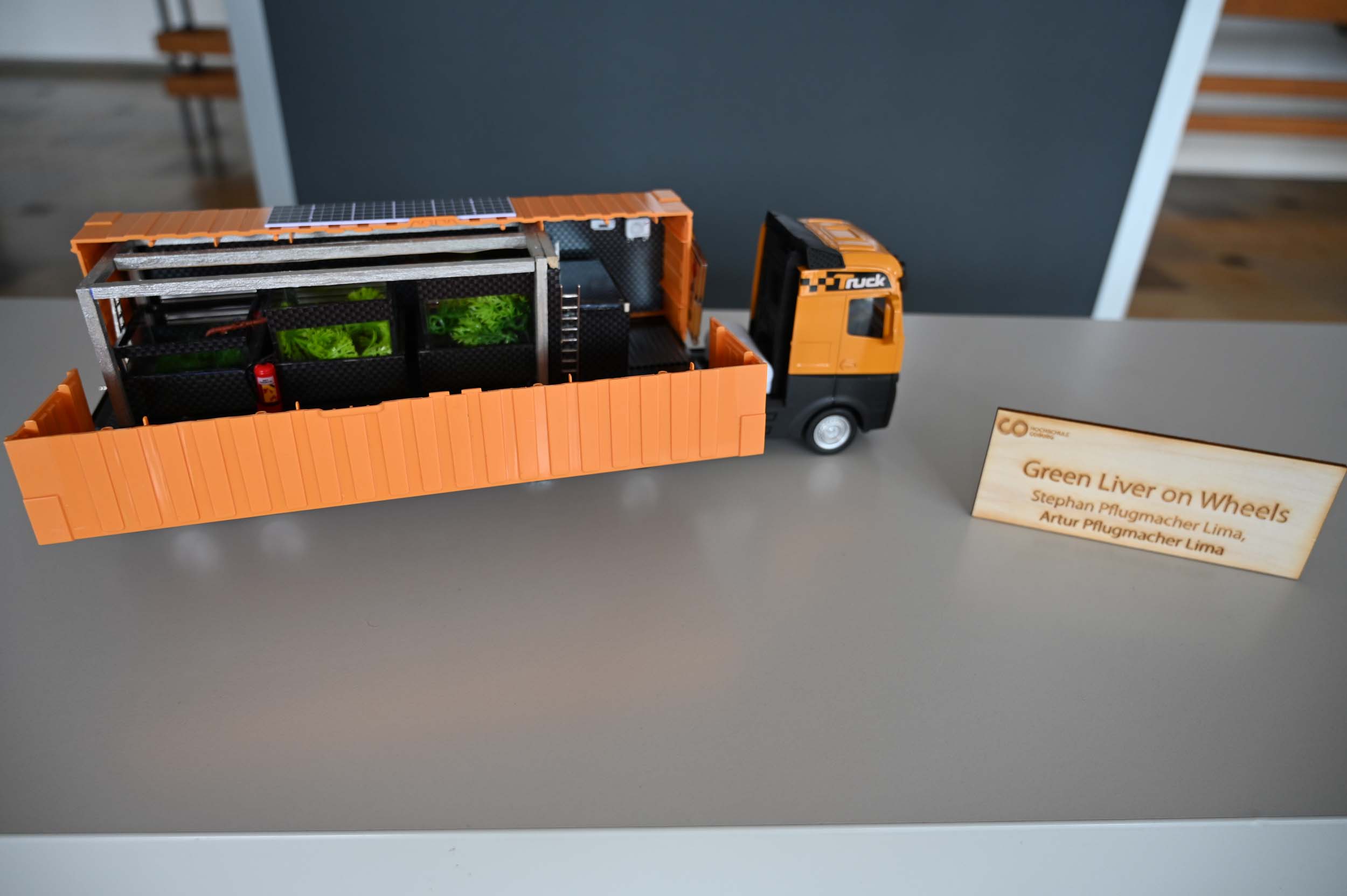 Ein Modell eines orangefarbenen Lastwagens, dessen aufgeschnittene Seite den Blick auf die Begrünung und die Ausrüstung im Inneren freigibt, steht auf einem grauen Tisch neben einem Schild mit der Aufschrift „Green Liver on Wheels“ und präsentiert innovative Lösungen für „Wasser retten“. at Hochschule Coburg