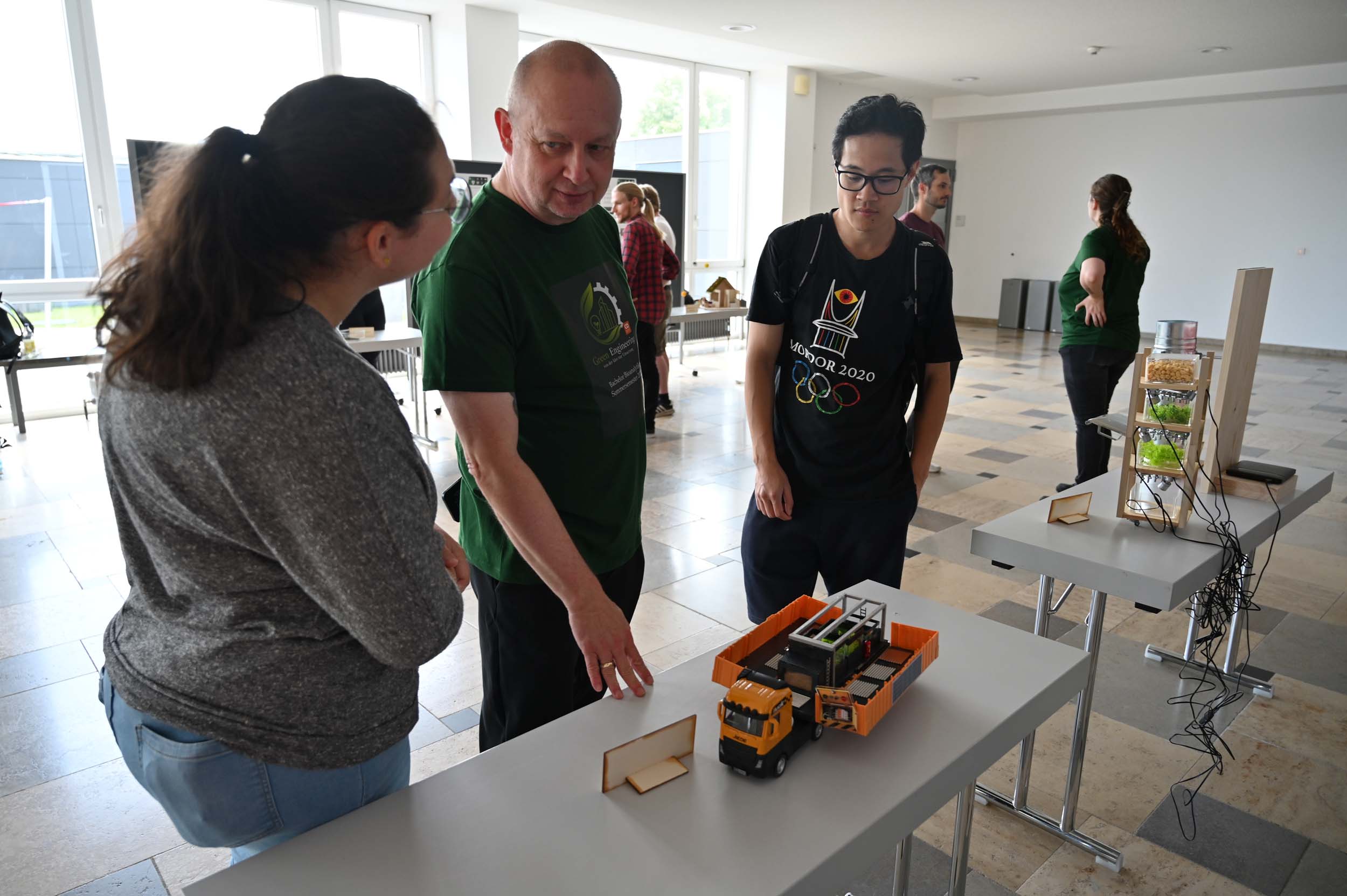 Drei Personen stehen um einen Tisch mit Spielzeug-Lkws und einem Modellgebäude herum und diskutieren oder begutachten die Gegenstände in einem hellen, modernen Raum, in dessen Hintergrund weitere Modelle und Menschen zu sehen sind – vielleicht im Rahmen einer Lehre oder eines Studiums, das sich mit innovativen Methoden zur Wasserrettung befasst. at Hochschule Coburg