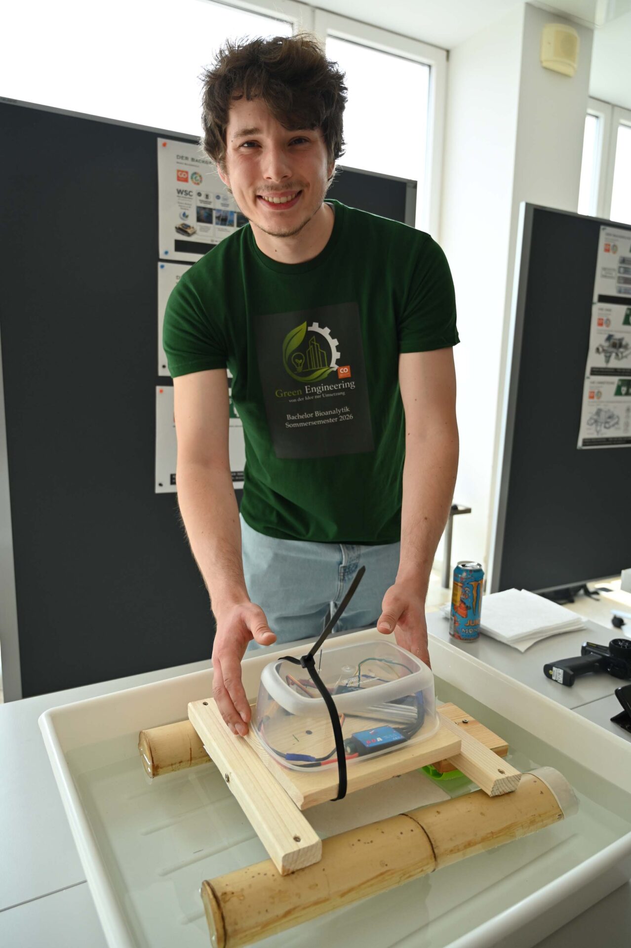 Ein lächelnder junger Mann in einem grünen Hemd demonstriert auf einer Wissenschaftsmesse ein kleines Floß aus Bambus und Holz mit elektronischer Ausstattung auf dem Wasser und präsentiert damit seinen innovativen Ansatz zum Thema „Wasser retten“, den er während seines Studiums entwickelt hat. at Hochschule Coburg