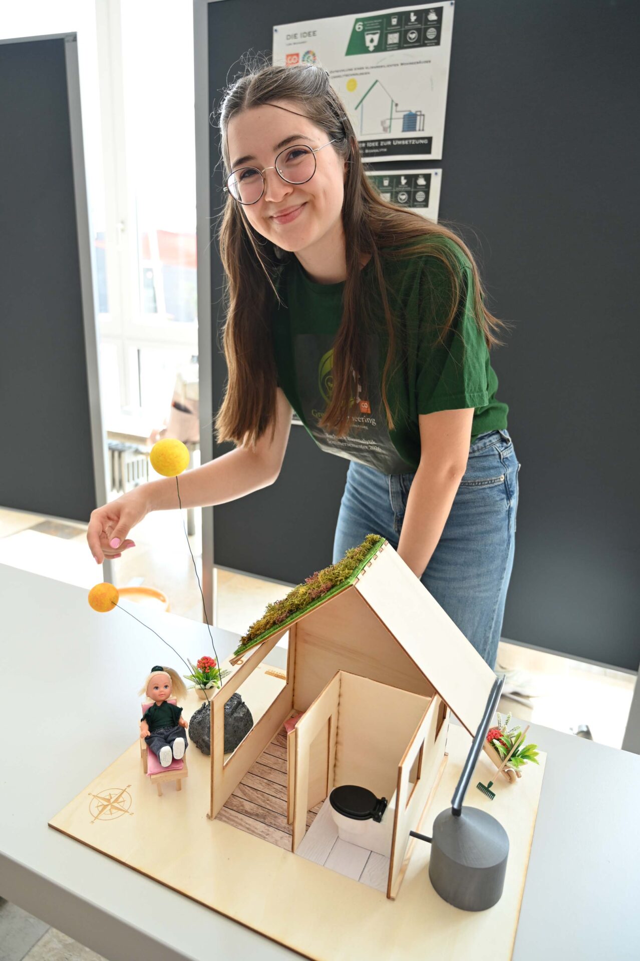 Eine lächelnde junge Frau steht neben einem Modellhaus und nimmt letzte Details vor – vielleicht im Rahmen ihres Studiums oder ihrer Ausbildung. Das Modell enthält eine kleine Puppe, Pflanzen und verschiedene Elemente, darunter auch solche, die auf kreative Weise veranschaulichen, wie man Wasser sparen kann. Es steht auf einem Tisch in einem Raum, im Hintergrund sind Plakate zu sehen. at Hochschule Coburg