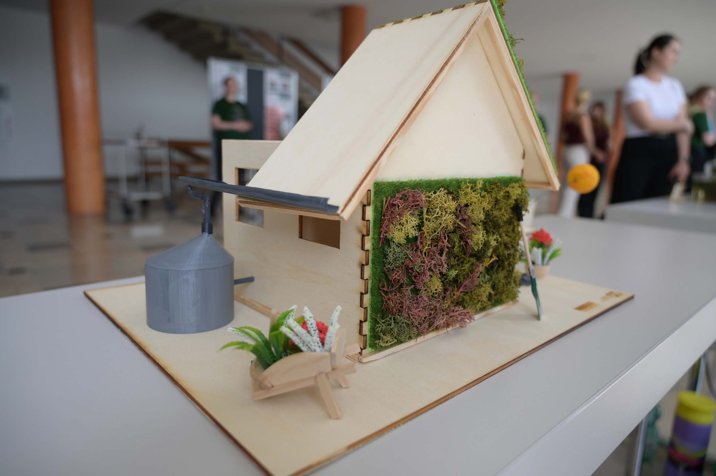 Ein kleines Holzhausmodell mit einer grünen Mooswand, einem Wassertank mit der Aufschrift „Wasser retten“ und einer Schubkarre auf einem Tisch. Im unscharfen Hintergrund sind Menschen und eine Treppe zu erkennen, was auf eine Lernumgebung hindeutet, die sich perfekt für Projekte im Rahmen von Lehre oder Studium zum Thema Nachhaltigkeit eignet. at Hochschule Coburg