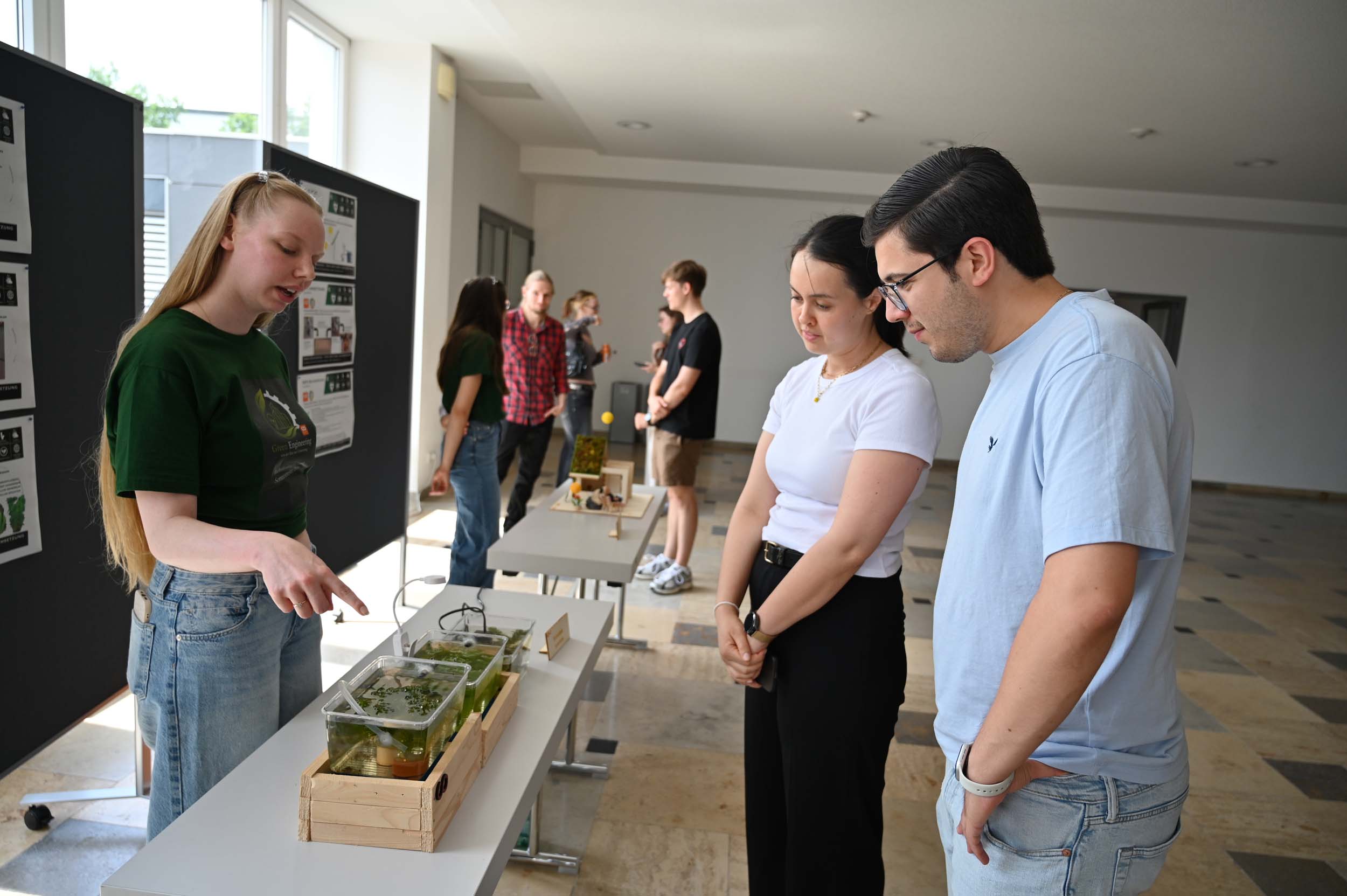 In einem hellen Raum erklärt eine Frau zwei Personen an einem Tisch anhand kleiner Pflanzen- und Wassertankmodelle die Bedeutung von „Wasser retten“, während andere im Hintergrund die Ausstellungsstücke betrachten. at Hochschule Coburg