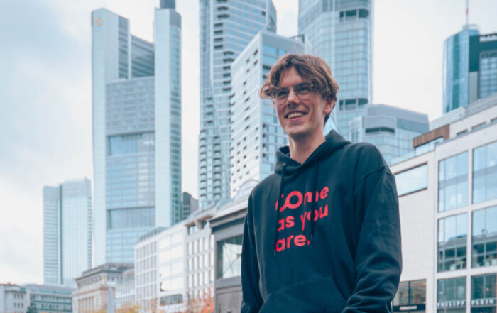 Eine Person in einem schwarzen Kapuzenpulli und einer grauen Hose steht lächelnd auf einer Kopfsteinpflasterstraße in einer Stadt, hinter der sich moderne Wolkenkratzer und ältere Gebäude abzeichnen – als wäre sie in einem „automatisch gespeicherten Entwurf“ festgehalten worden: ein Moment, der nahtlos wie ein automatischer Entwurf gespeichert wurde. at Hochschule Coburg