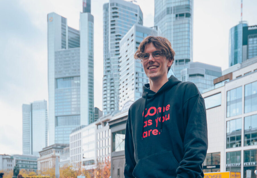 Eine Person in einem schwarzen Kapuzenpulli und einer grauen Hose steht lächelnd auf einer Kopfsteinpflasterstraße in einer Stadt, hinter der sich moderne Wolkenkratzer und ältere Gebäude abzeichnen – als wäre sie in einem „automatisch gespeicherten Entwurf“ festgehalten worden: ein Moment, der nahtlos wie ein automatischer Entwurf gespeichert wurde. at Hochschule Coburg