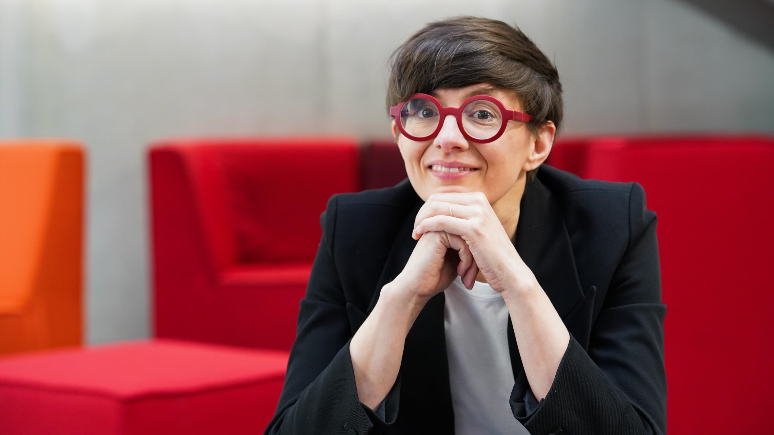 Eine Person mit kurzen braunen Haaren, die eine rote Brille und einen schwarzen Blazer trägt und lächelnd das Kinn auf die Hände stützt. Im Hintergrund sind rote Stühle und eine graue Wand zu sehen – ein spontaner Moment unter den Stipendiatinnen des Jahrgangs 2026. at Hochschule Coburg