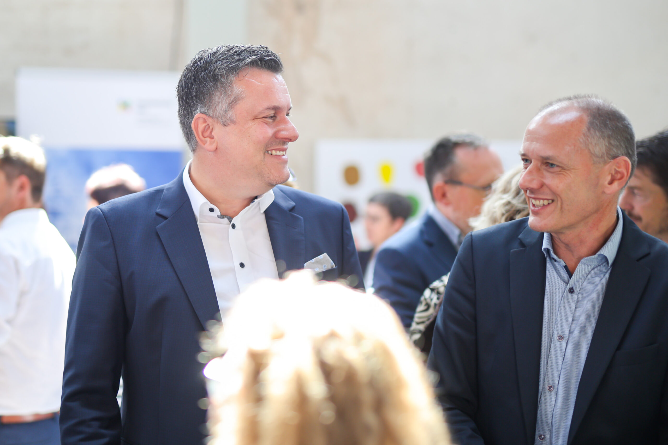Zwei Männer in Anzügen lächeln und unterhalten sich bei einer Veranstaltung in Kronach, bei der „zehn Jahre Hochschulstandort Kronach“ gefeiert wird. Der hell beleuchtete, unscharfe Hintergrund vermittelt eine lebendige und professionelle Atmosphäre. at Hochschule Coburg