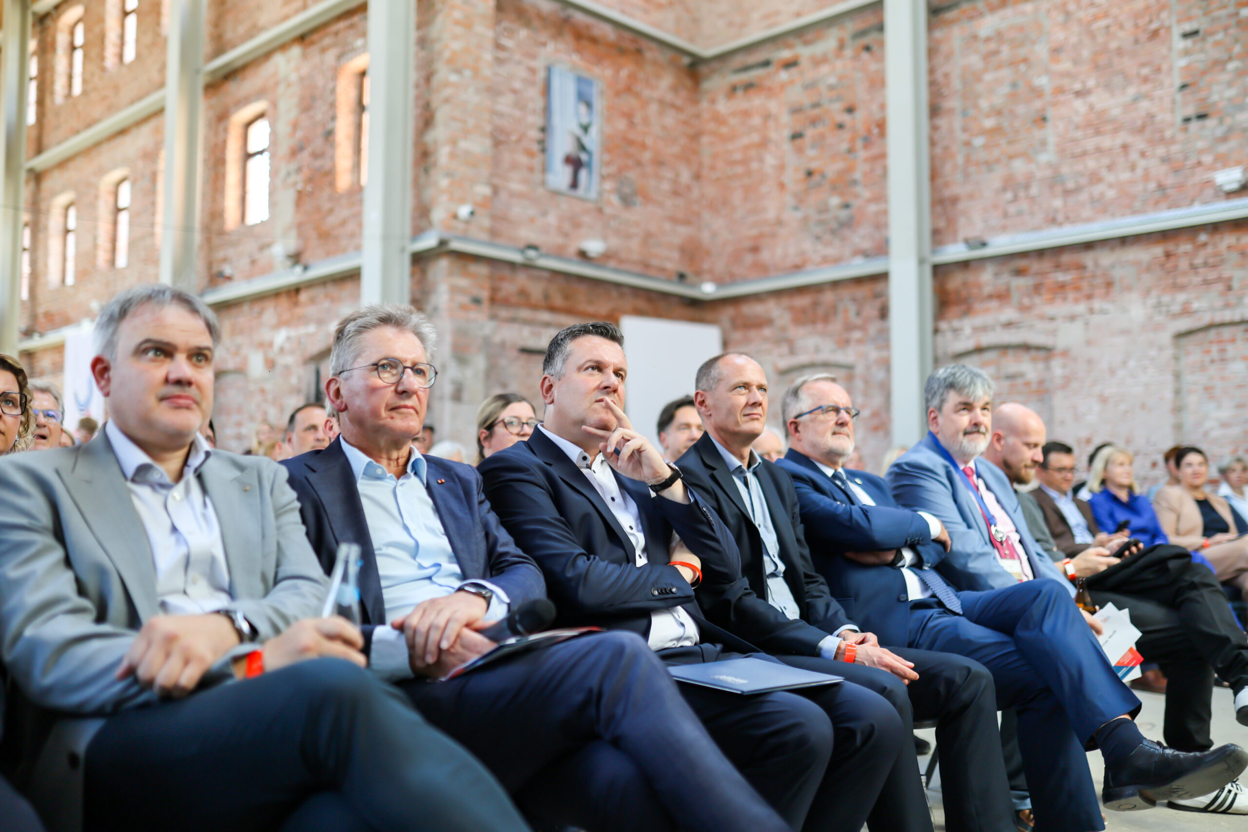 Eine Gruppe von Männern in Anzügen sitzt in der ersten Reihe bei einer Veranstaltung in einem Saal mit unverputzten Ziegelwänden am Hochschulstandort Kronach und hört aufmerksam einer Präsentation zu. Im Hintergrund sind weitere Teilnehmer zu sehen. at Hochschule Coburg