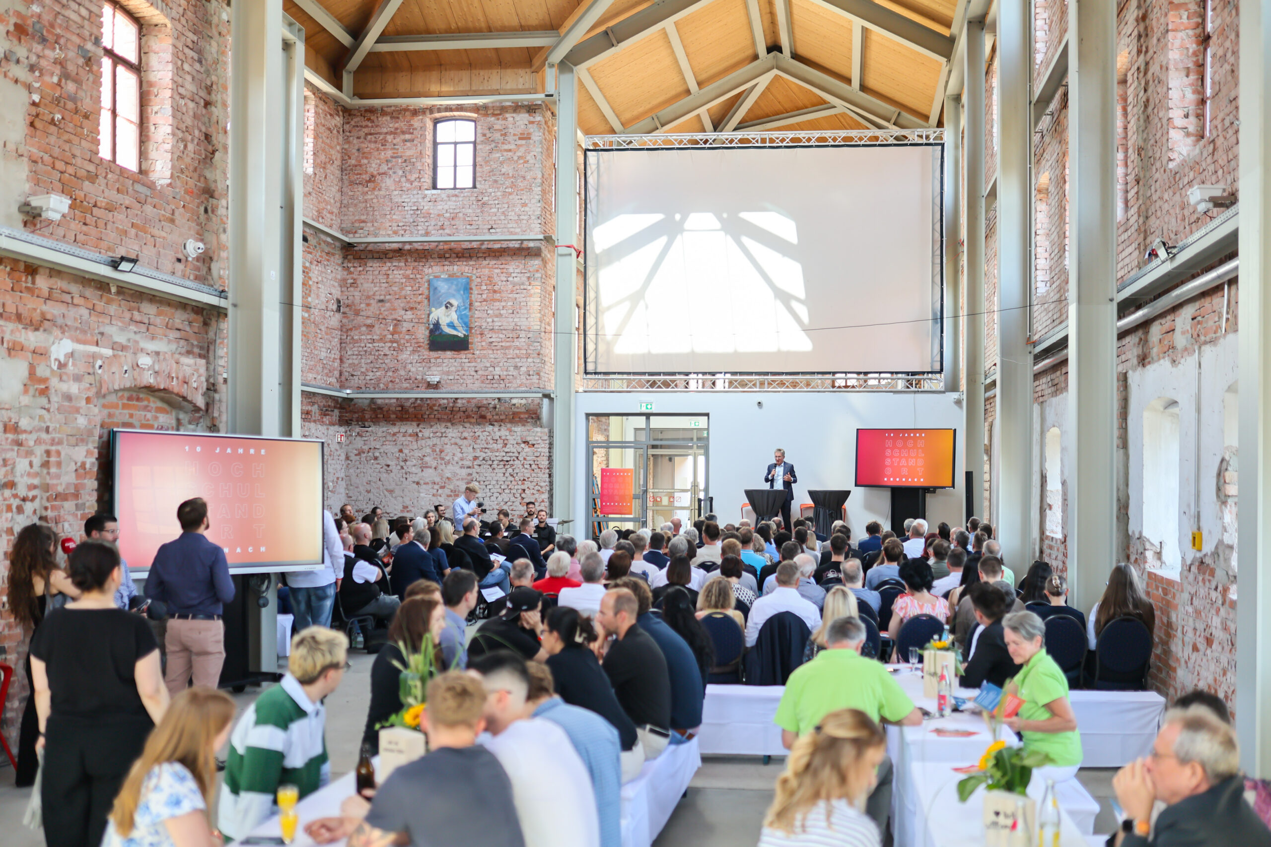 Eine große Menschenmenge nimmt an einer Jubiläumsveranstaltung in einem geräumigen Saal mit unverputzten Ziegelwänden, hohen Decken und großen Fenstern teil. Der Redner auf der Bühne feiert „zehn Jahre Hochschulstandort Kronach“, wobei Bildmaterial auf Bildschirmen und mit einem Projektor gezeigt wird. at Hochschule Coburg