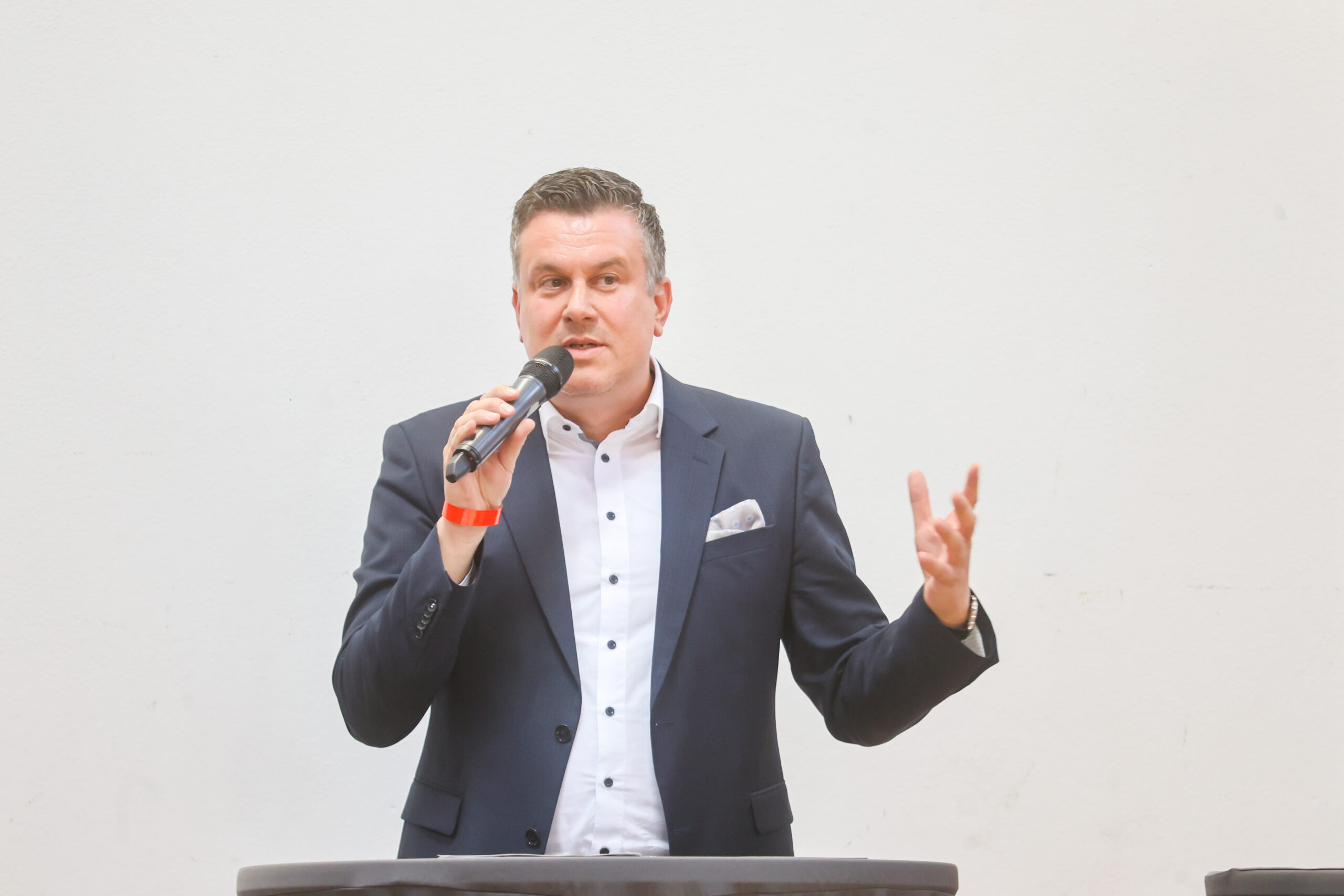 Ein Mann in einem dunklen Anzug und einem weißen Hemd spricht vor einem Rednerpult in ein Mikrofon, gestikuliert dabei vor einem schlichten weißen Hintergrund und feiert das zehnjährige Jubiläum des Hochschulstandorts Kronach. at Hochschule Coburg