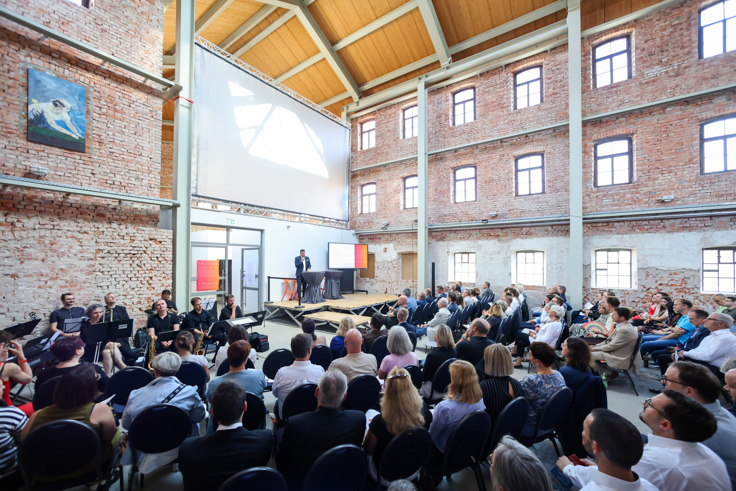 Ein Redner hält auf der Bühne vor einem sitzenden Publikum in einem geräumigen Saal im Industriestil am Hochschulstandort Kronach eine Rede; der Saal verfügt über unverputzte Ziegelwände, hohe Fenster und eine große Leinwand hinter dem Redner. Links sitzt ein kleines Orchester. at Hochschule Coburg
