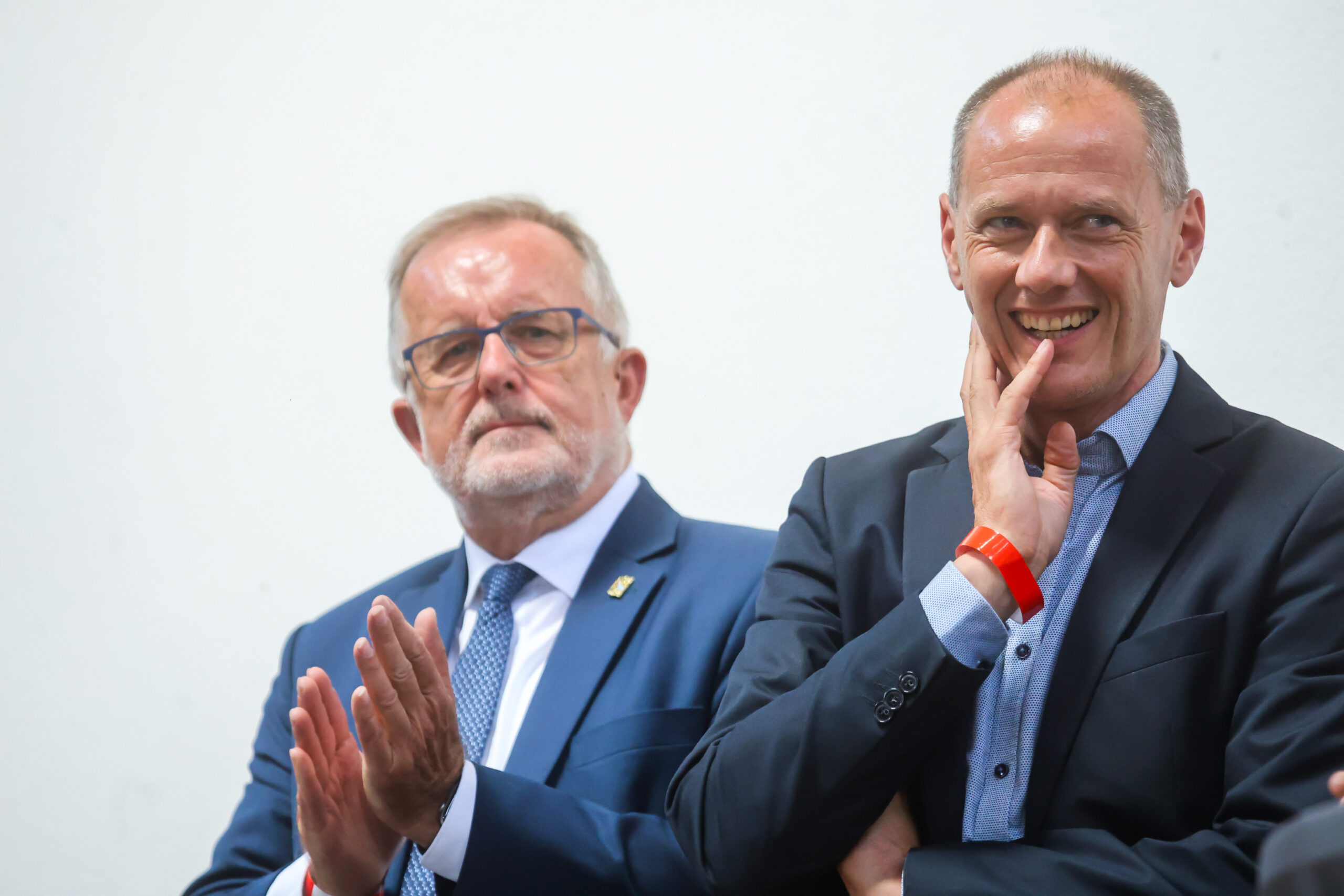 Zwei Männer in Anzügen stehen vor einem schlichten weißen Hintergrund. Der Mann links klatscht mit ernstem Gesichtsausdruck, während der Mann rechts lächelt und sich an die Wange fasst – anlässlich des zehnjährigen Jubiläums des Hochschulstandorts Kronach. at Hochschule Coburg
