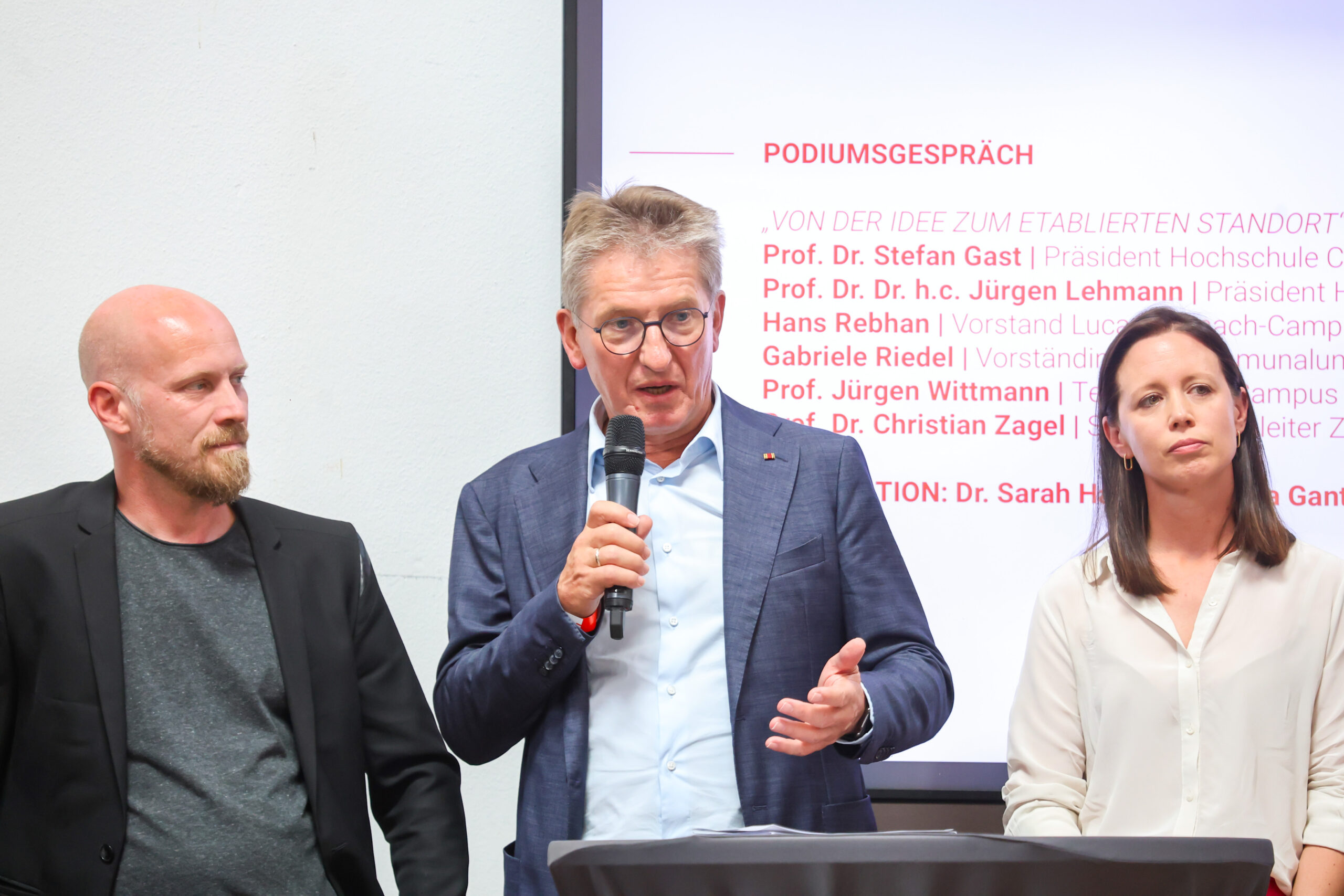 Drei Personen stehen während einer Podiumsdiskussion am Hochschulstandort Kronach vor einer Leinwand. Der Mann in der Mitte spricht in ein Mikrofon, während die beiden anderen, die das zehnjährige Jubiläum feiern, aufmerksam zuhören. at Hochschule Coburg