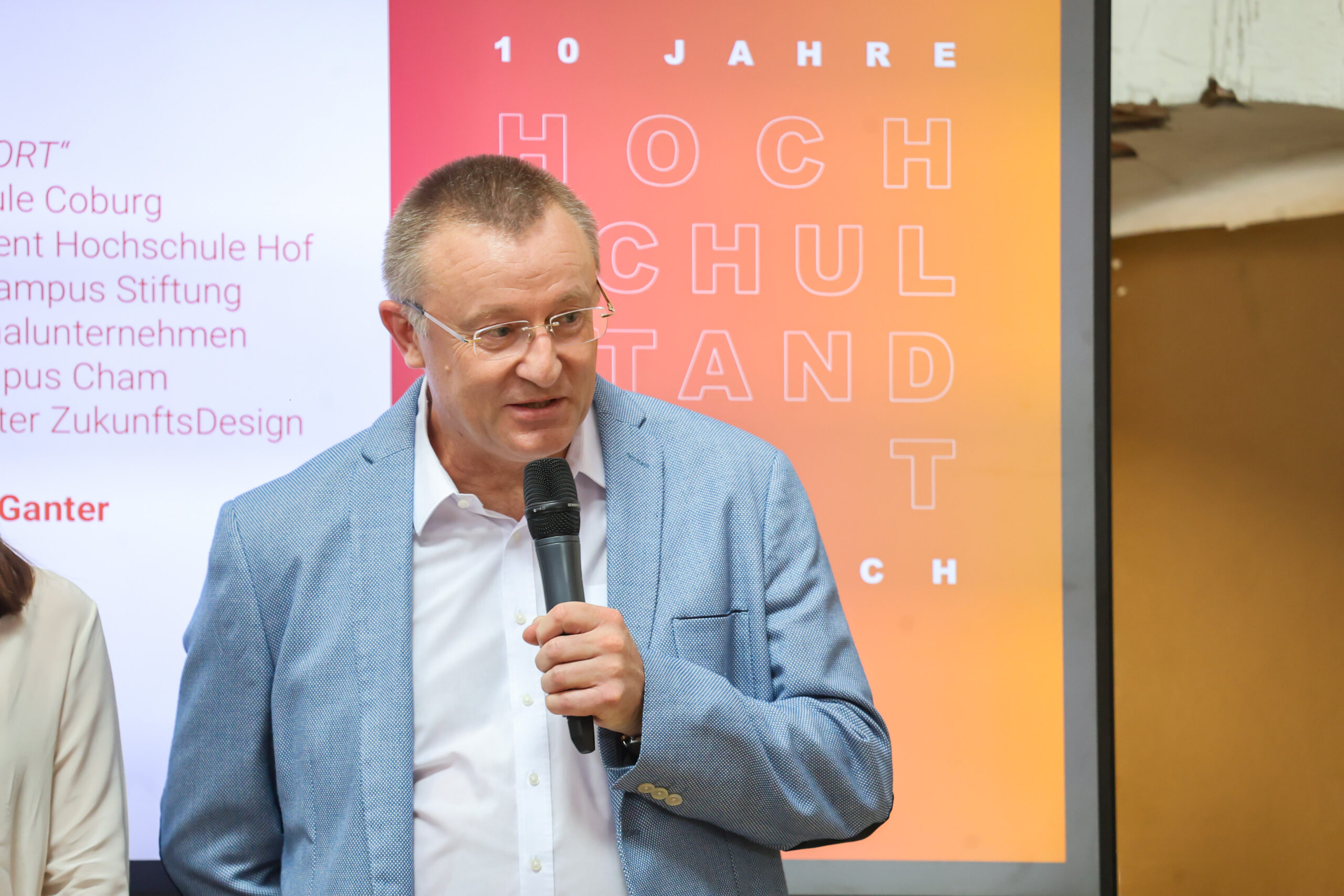 Ein Mann in einem hellblauen Blazer hält ein Mikrofon in der Hand und spricht. Hinter ihm ist auf einer Leinwand mit einem orange-rosa Farbverlauf ein deutscher Text zu sehen, der das 10-jährige Jubiläum des Hochschulstandorts Kronach feiert. at Hochschule Coburg