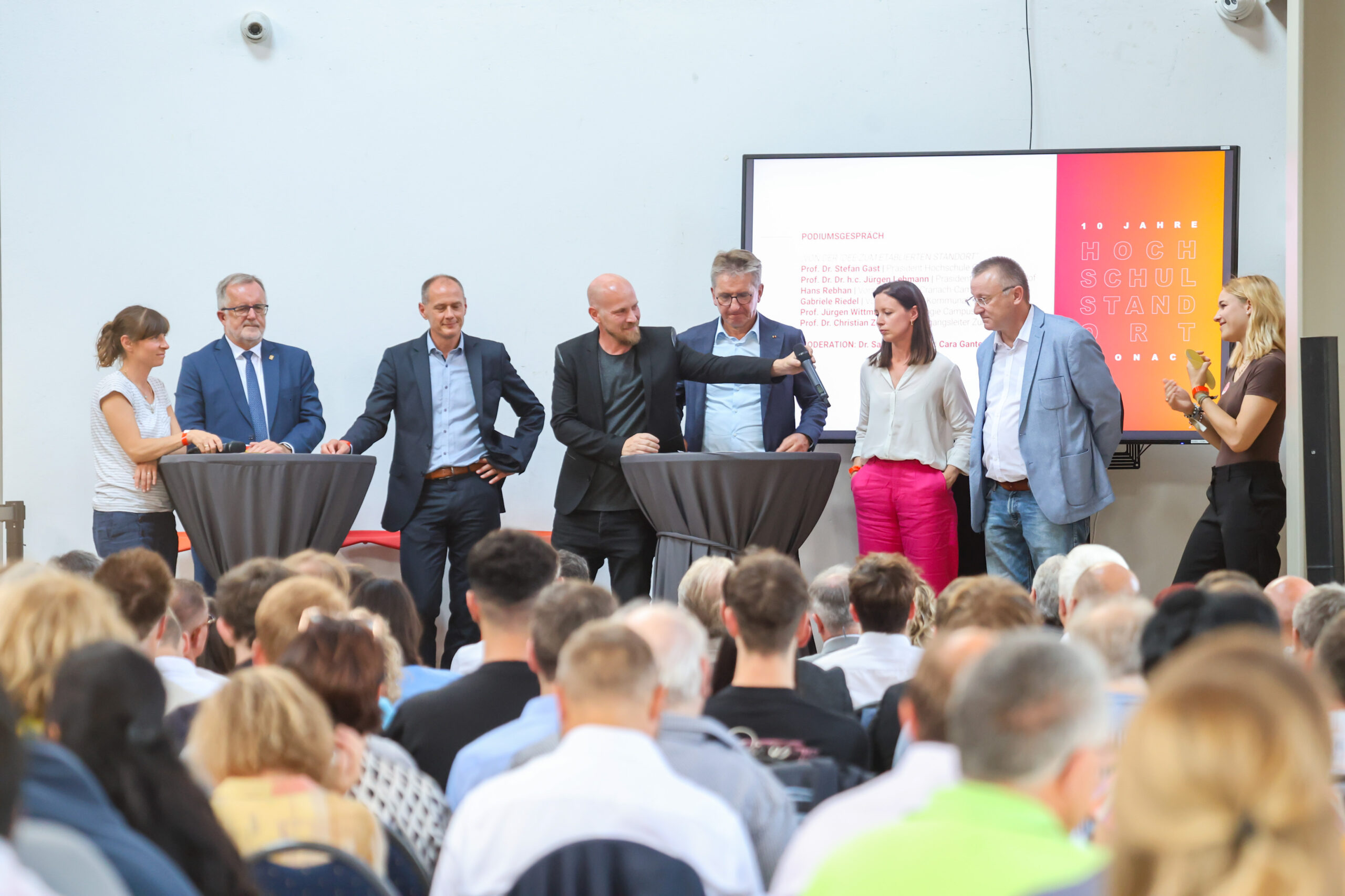 Sieben Personen stehen auf einer Bühne hinter Cocktailtischen und sprechen zu den Zuhörern. Im Hintergrund der Podiumsdiskussion oder Präsentation zum Thema „Zehn Jahre Hochschulstandort Kronach“ ist eine große Leinwand mit farbenfrohem Text zu sehen. at Hochschule Coburg