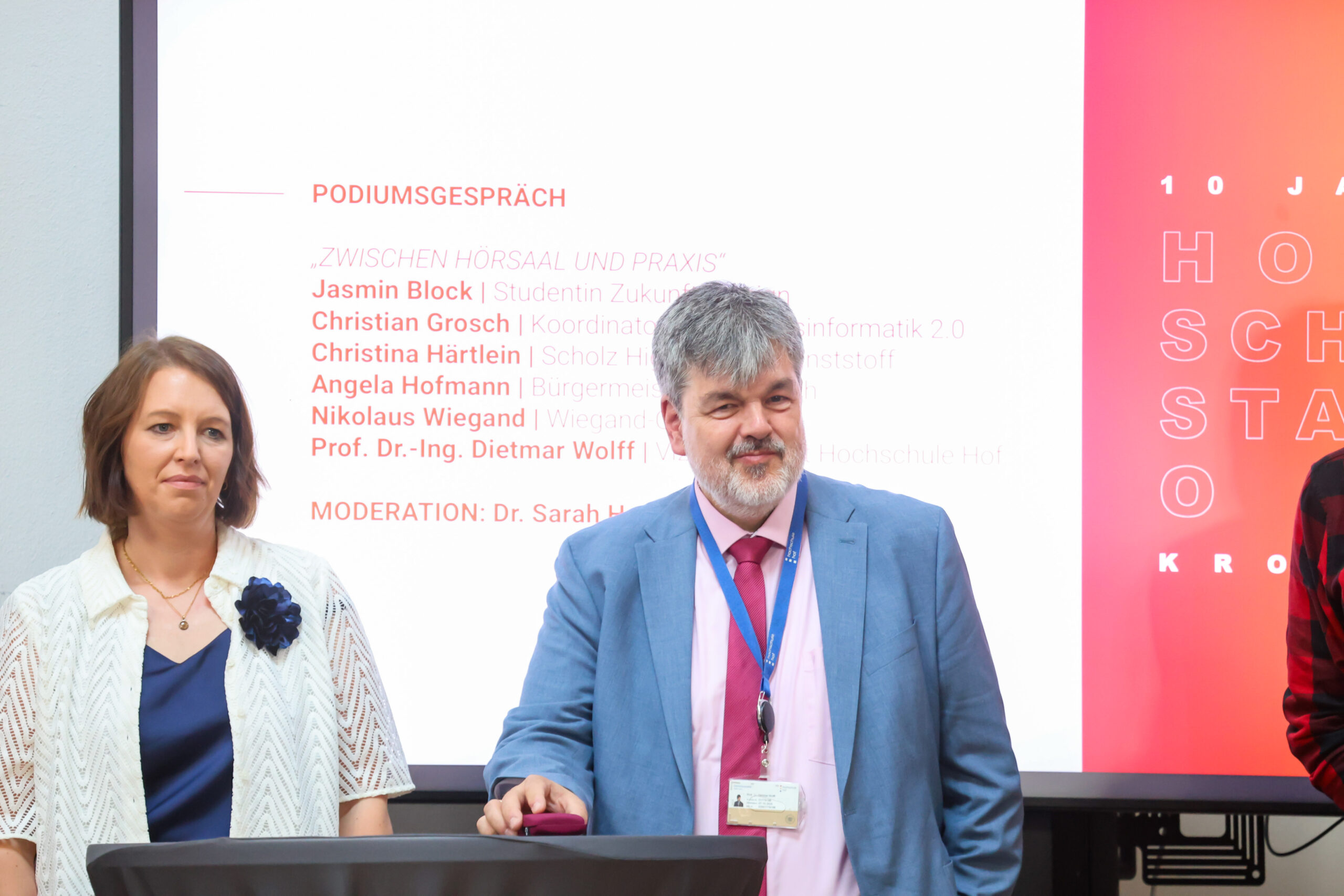 Am Hochschulstandort Kronach stehen eine Frau und ein Mann vor einer Leinwand, auf der das Programm einer Podiumsdiskussion auf Deutsch angezeigt wird. Der Mann trägt einen Anzug und ein Umhängeband, während die Frau eine leichte Jacke und eine Brosche trägt. Beide blicken nach vorne. at Hochschule Coburg