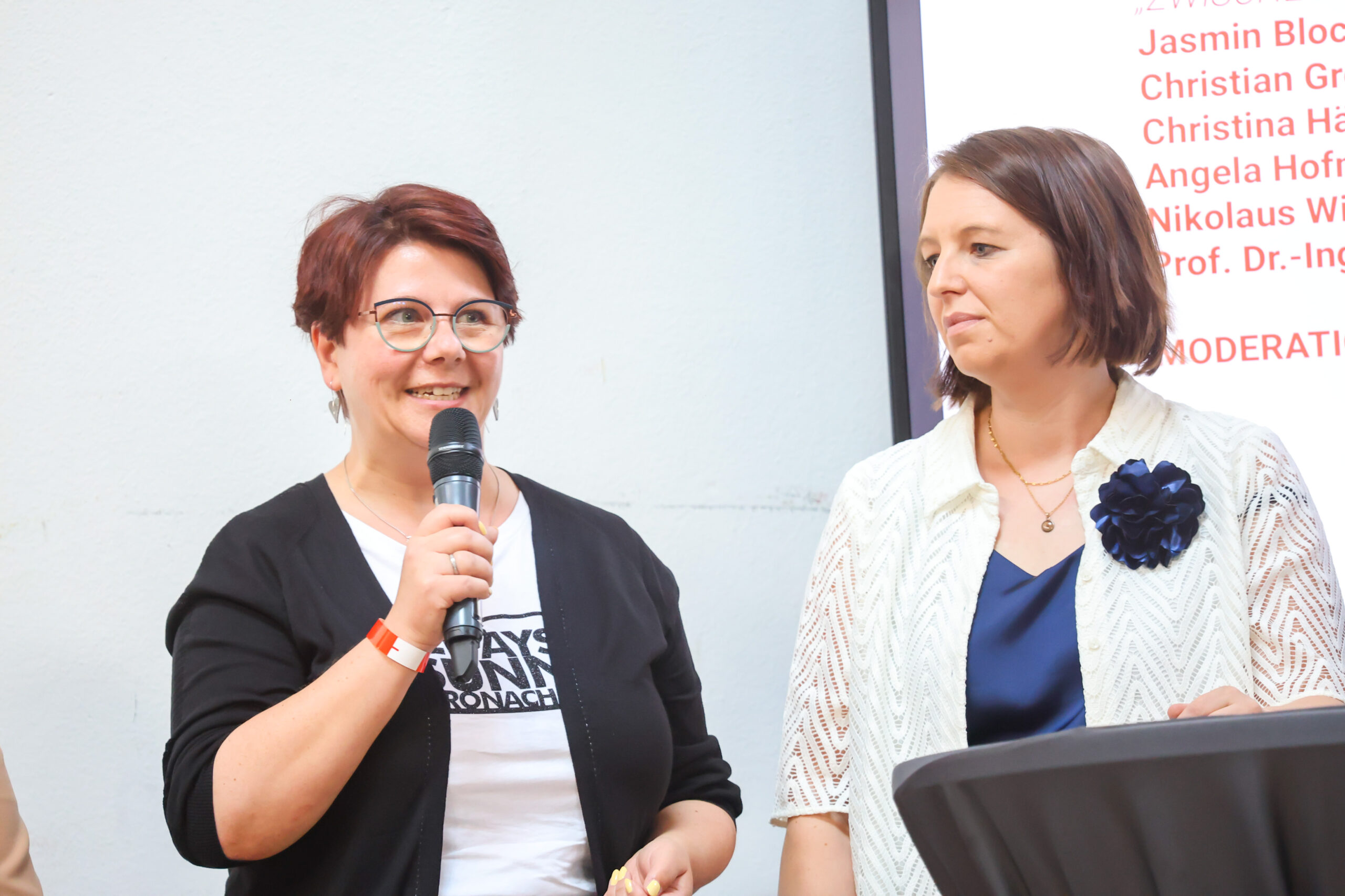 Zwei Frauen stehen bei einer Veranstaltung in Kronach nebeneinander. Die Frau links, die lächelt, hält ein Mikrofon in der Hand und trägt eine Brille sowie ein weißes Hemd. Die Frau rechts, die eine weiße Jacke trägt, schaut sie an. Hinter ihnen ist eine Leinwand mit einem Text zum Thema „Hochschulstandort“ zu sehen. at Hochschule Coburg