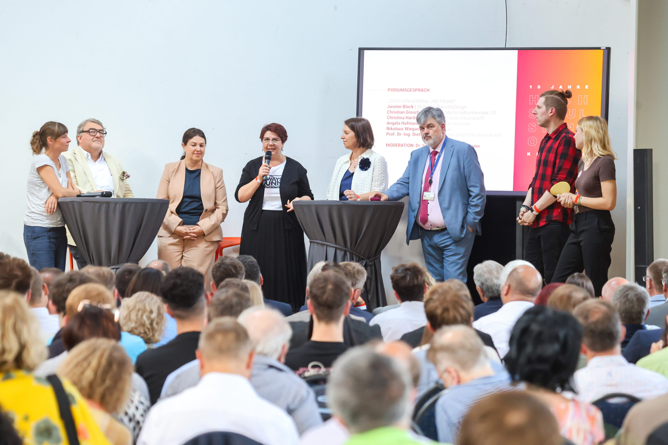 Eine Gruppe von acht Personen steht auf einer Bühne in Kronach und spricht zu den Zuhörern. Einige stehen hinter hohen Tischen; eine Person hält ein Mikrofon in der Hand. Im Hintergrund ist eine Präsentationswand mit einem Text zum Thema „Zehn Jahre Hochschulstandort Kronach“ zu sehen. at Hochschule Coburg