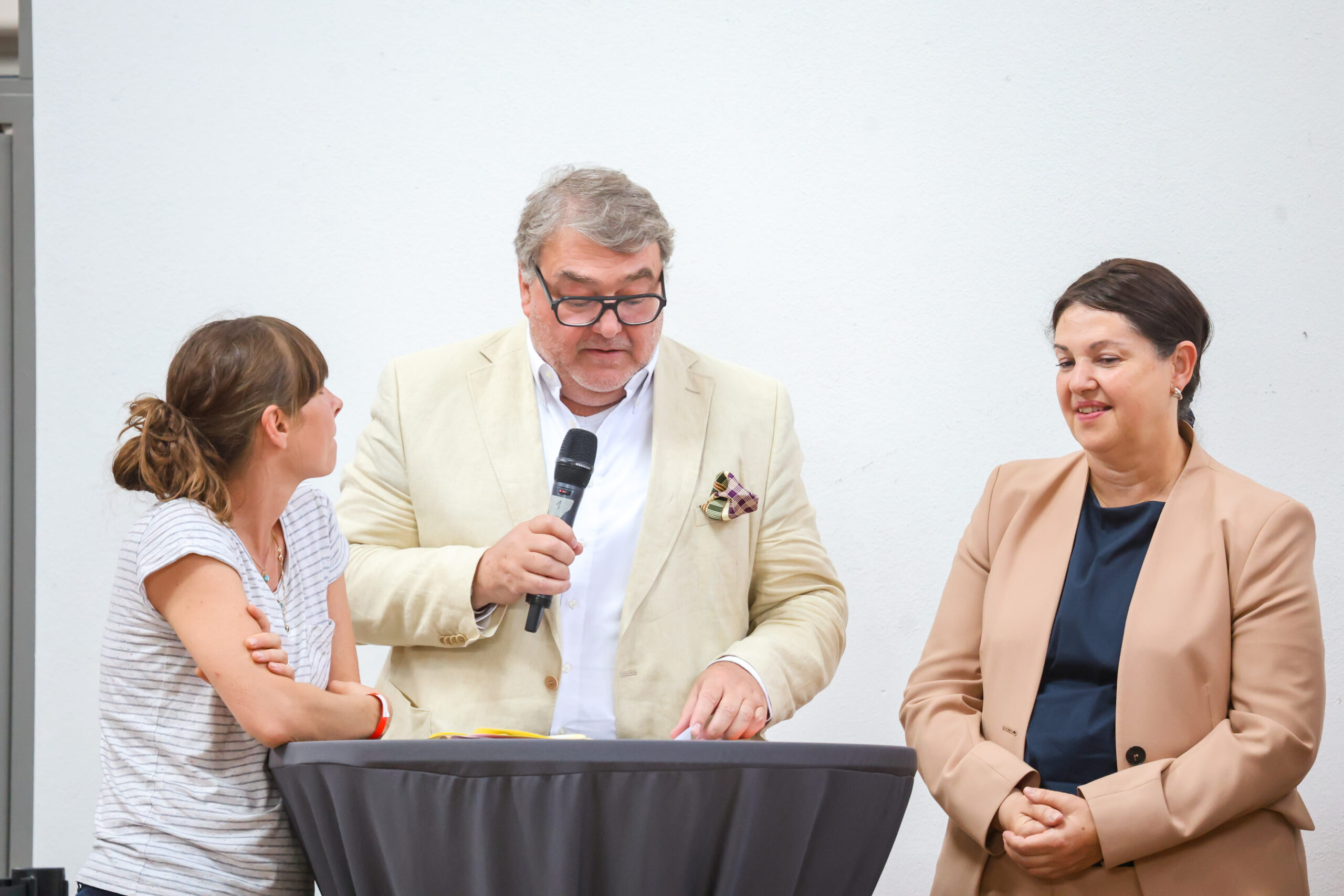 Am Hochschulstandort Kronach stehen drei Personen um einen runden, gedeckten Tisch herum; ein Mann in einem beigen Anzug hält ein Mikrofon in der Hand und spricht, während zwei Frauen – eine in einem gestreiften Hemd und eine in einem hellbraunen Blazer – aufmerksam zuhören. at Hochschule Coburg
