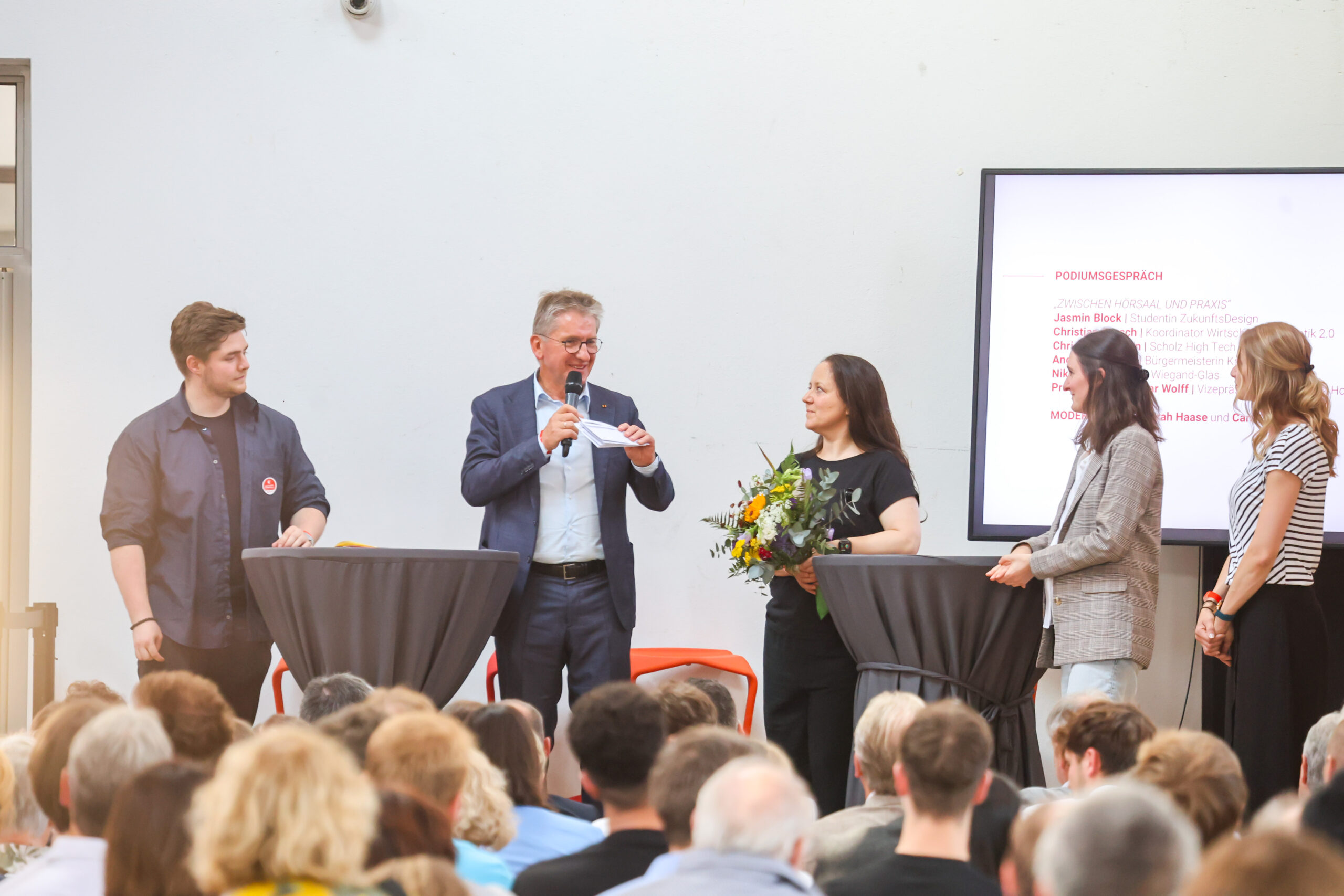 Eine Gruppe von fünf Personen, darunter eine, die einen Blumenstrauß in der Hand hält, spricht vor Publikum in Kronach. Ein Mann spricht ins Mikrofon, und im Hintergrund ist auf einer großen Leinwand der Schriftzug „zehn Jahre Hochschulstandort Kronach“ zu sehen. at Hochschule Coburg
