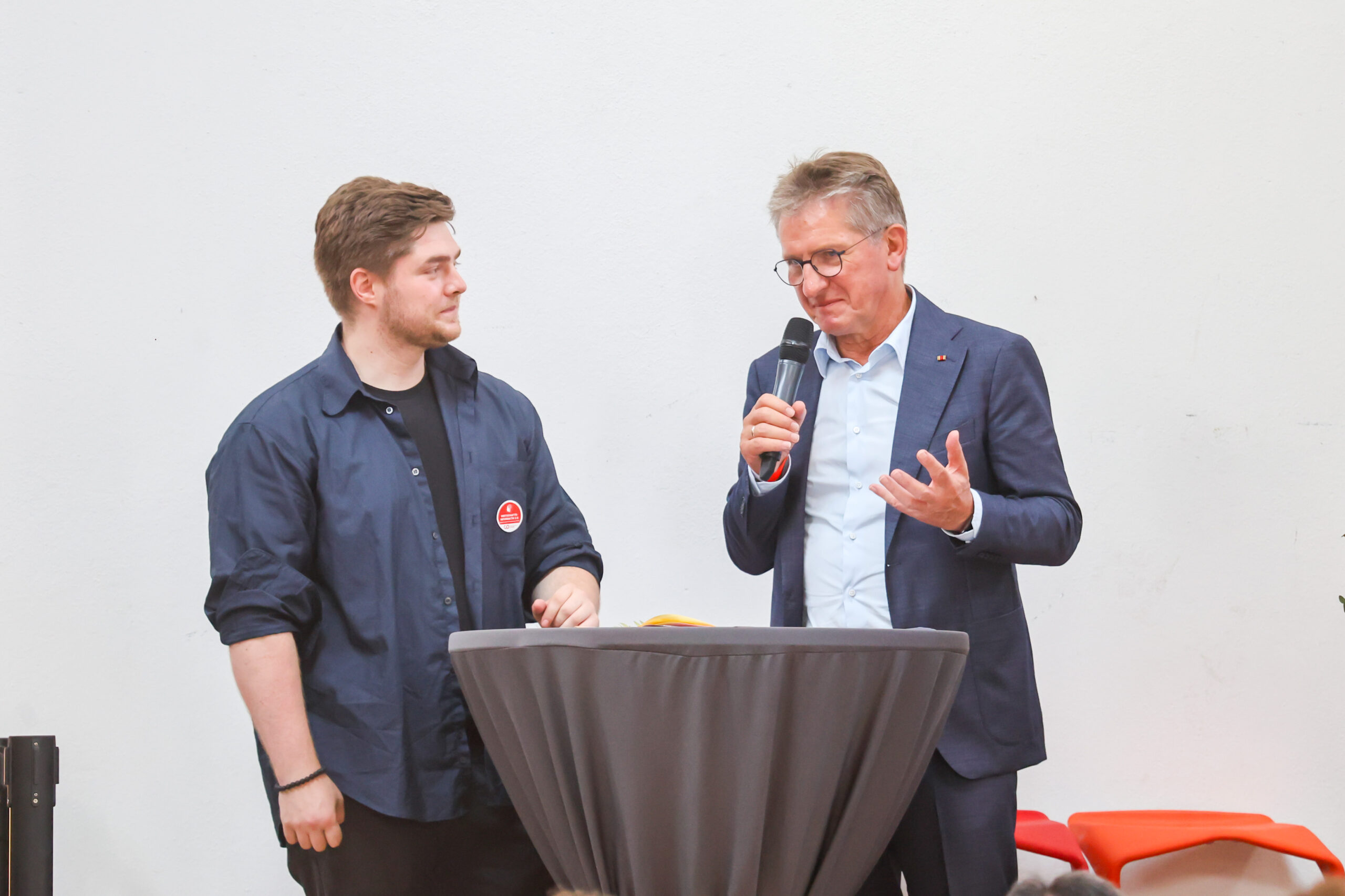 Zwei Männer stehen an einem runden Tisch; der eine, in einem dunklen Hemd, blickt den anderen an, der einen Anzug und eine Brille trägt und in ein Mikrofon spricht. Vor einem schlichten weißen Hintergrund scheinen sie über „zehn Jahre Hochschulstandort Kronach“ zu sprechen. at Hochschule Coburg