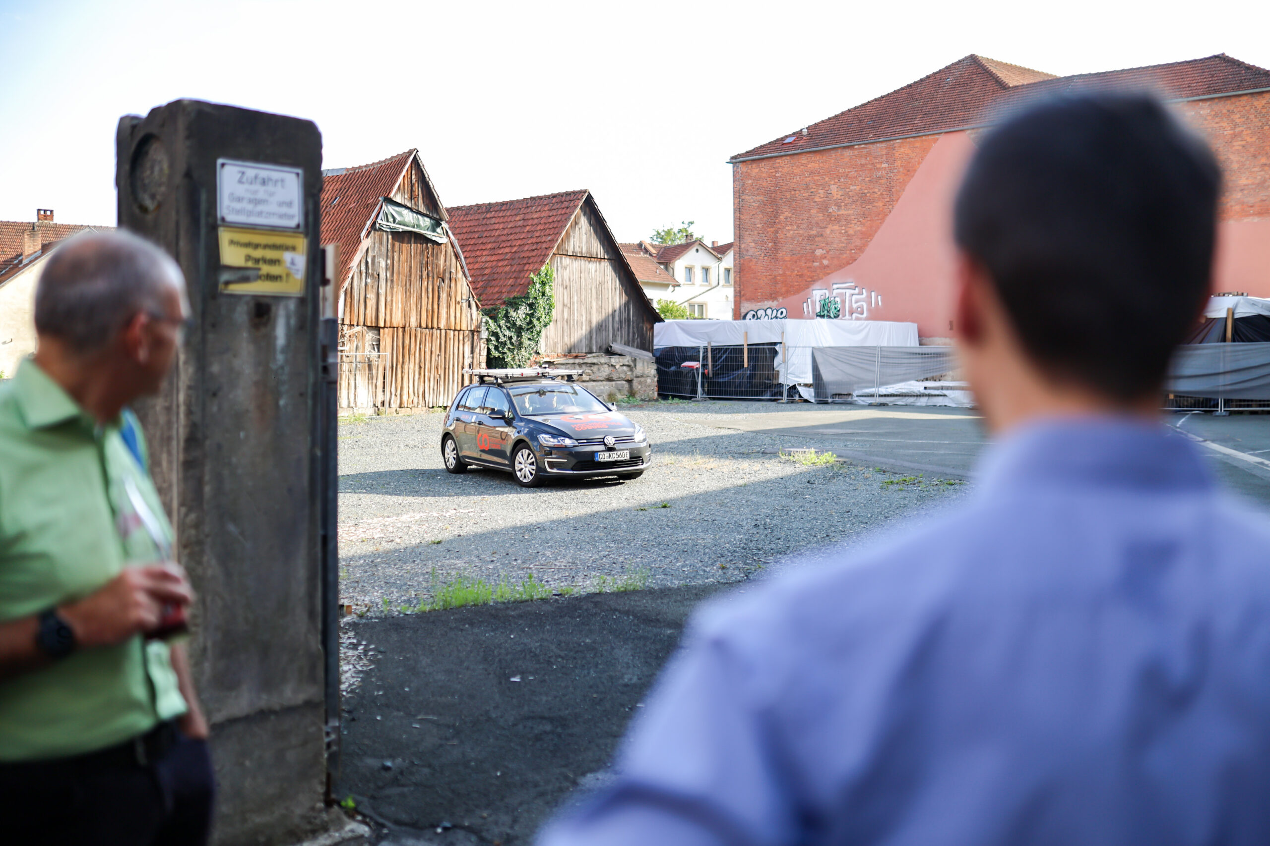 Im Vordergrund stehen zwei Männer und betrachten ein kleines schwarzes Auto, das auf einem Schotterplatz geparkt ist; im Hintergrund sind Holzscheunen und ein rotes Backsteingebäude zu sehen – ein Schnappschuss aus dem Alltag in Kronach, in der Nähe des Hochschulstandorts Kronach. at Hochschule Coburg