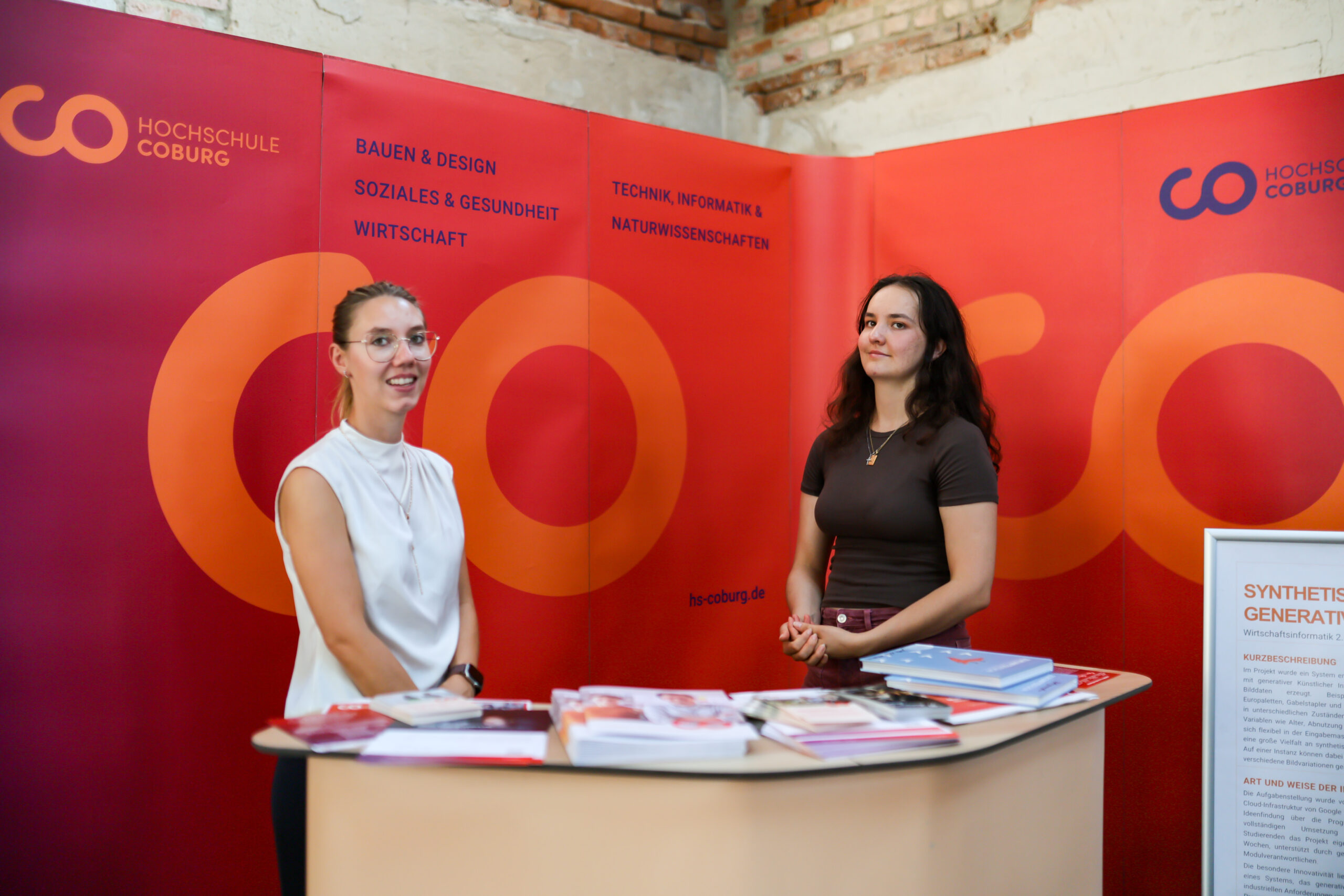 Zwei Frauen stehen hinter einem Informationsstand bei einer Veranstaltung der Hochschule Coburg in Kronach. Im Hintergrund sind das Logo der Hochschule und die Namen der Fachbereiche auf Deutsch zu sehen. Es liegen Broschüren und Flyer aus, die das Jubiläum des Hochschulstandorts Kronach feiern. at Hochschule Coburg