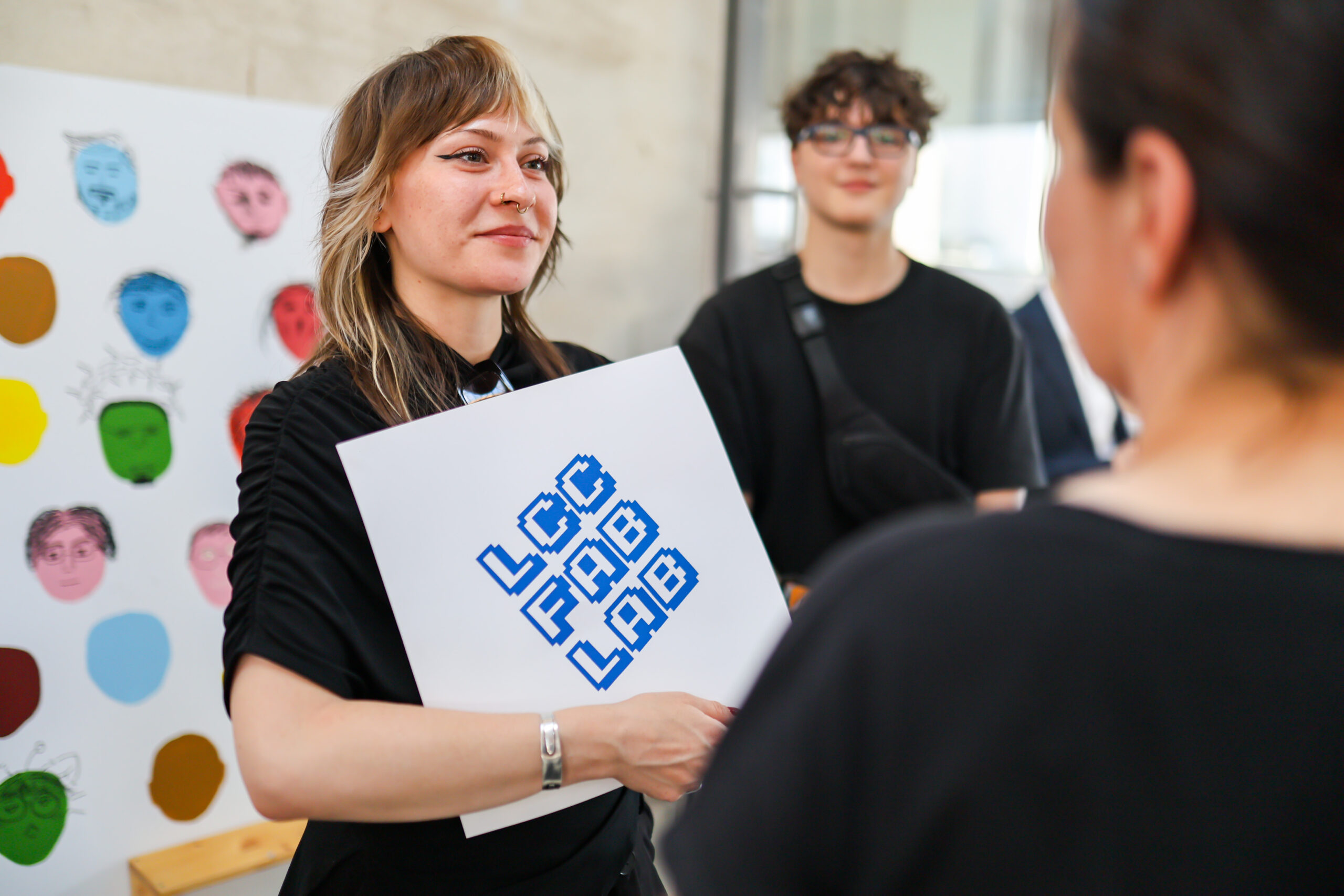 Eine Frau, die ein Schild mit der Aufschrift „LCC FAB LAB“ in der Hand hält, steht am Hochschulstandort Kronach vor einem bunten Plakat mit illustrierten Gesichtern und spricht mit jemandem, der nicht im Bild zu sehen ist. Im Hintergrund ist eine weitere Person unscharf zu erkennen. at Hochschule Coburg