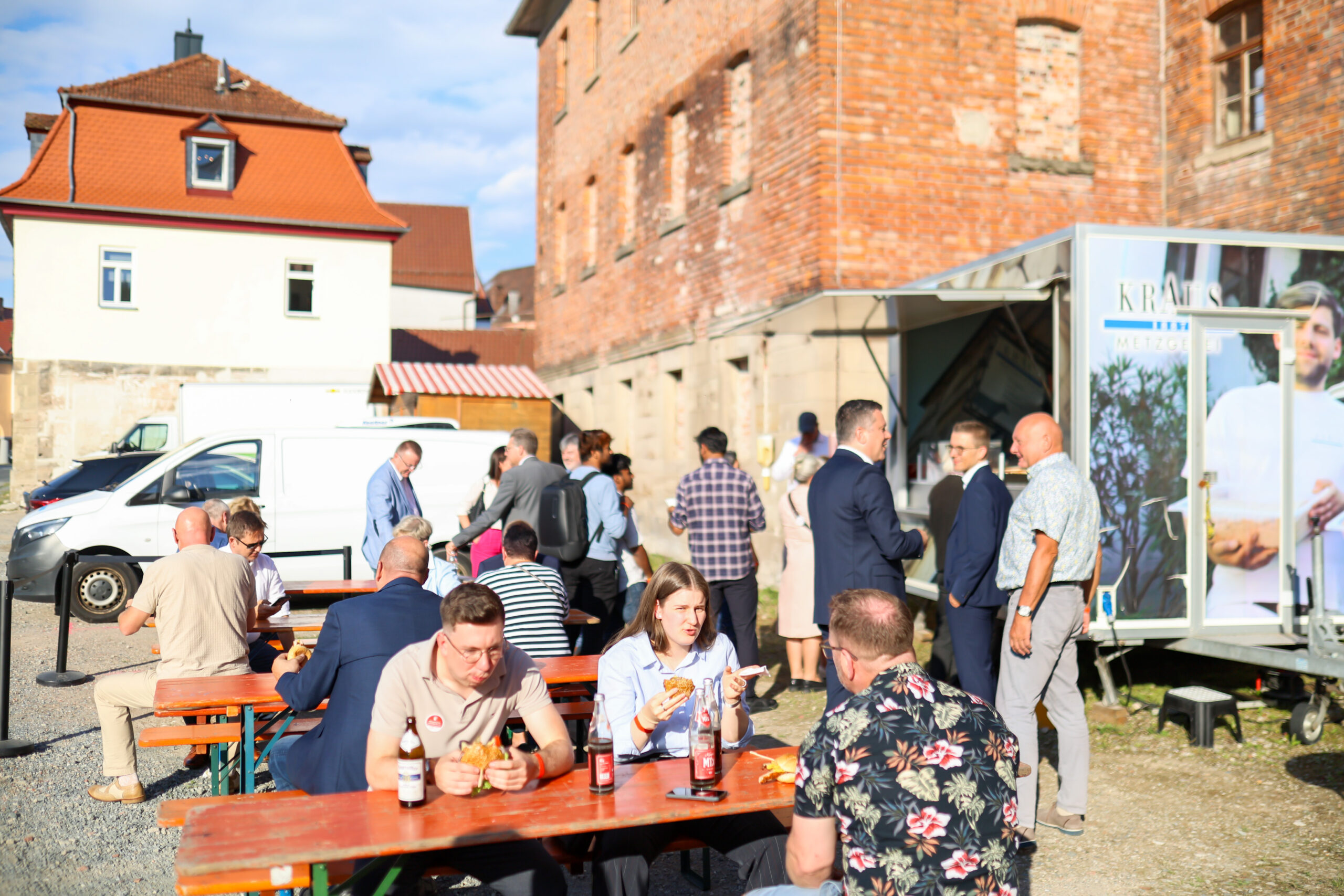 An einem sonnigen Tag versammeln sich die Menschen im Freien an Picknicktischen und stehen in Gruppen beieinander, unterhalten sich und essen in der Nähe von Backsteingebäuden und eines weißen Lieferwagens – so entsteht eine ungezwungene, gesellige Atmosphäre, während „Zehn Jahre Hochschulstandort Kronach“ gefeiert wird. at Hochschule Coburg