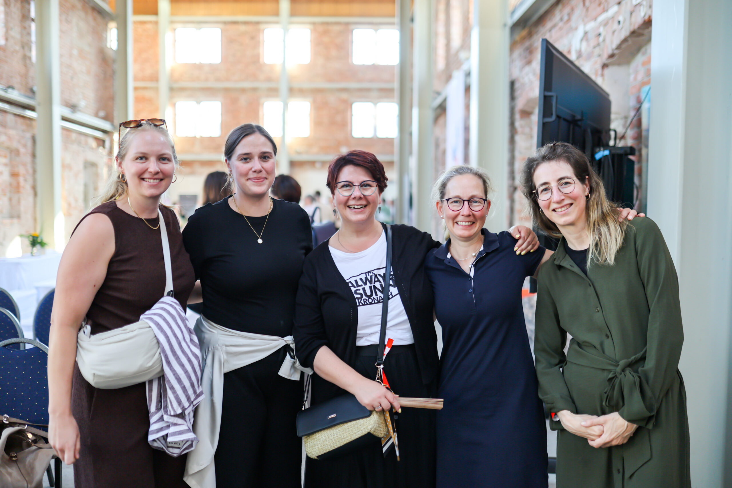 Fünf Frauen stehen eng beieinander und lächeln in die Kamera – in einem hellen Raum im Industrial-Stil am Hochschulstandort Kronach, mit unverputzten Ziegelwänden, großen Fenstern sowie Tischen und Stühlen im Hintergrund. at Hochschule Coburg