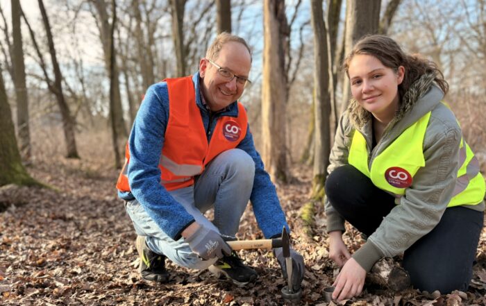 Zwei Personen in Warnwesten knien auf dem Waldboden und pflanzen gemeinsam etwas in den Boden. Sie sind von blattlosen Bäumen umgeben, und Sonnenlicht fällt durch die Äste. Beide wirken konzentriert und vertieft in ihre Tätigkeit, was darauf hindeutet, dass sie möglicherweise an Forschungsarbeiten zu Umweltauswirkungen beteiligt sind oder sogar das Vorkommen von Mikroplastik in Waldökosystemen untersuchen. at Hochschule Coburg
