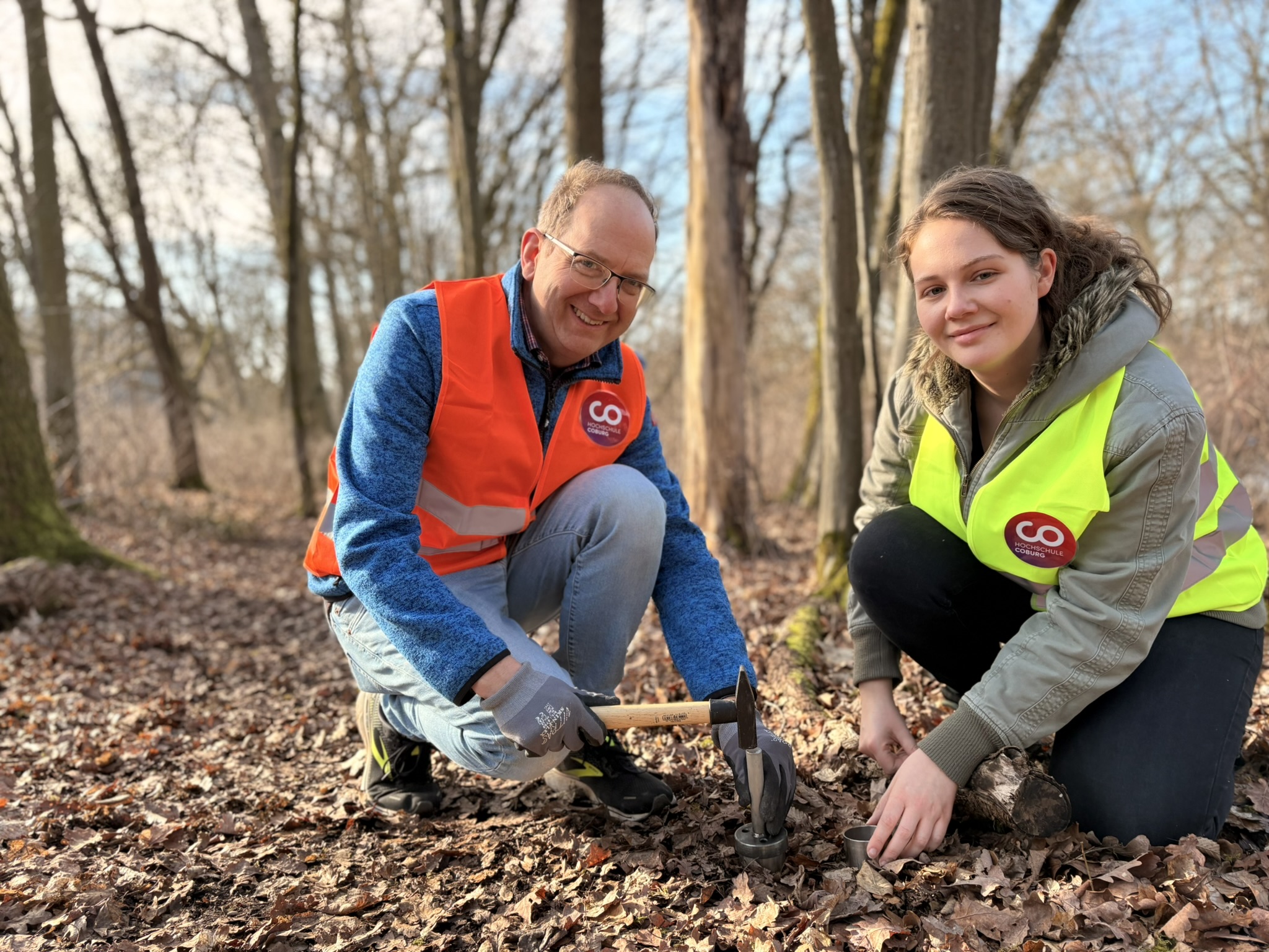 Zwei Personen in Warnwesten knien auf dem Waldboden und pflanzen gemeinsam etwas in den Boden. Sie sind von blattlosen Bäumen umgeben, und Sonnenlicht fällt durch die Äste. Beide wirken konzentriert und vertieft in ihre Tätigkeit, was darauf hindeutet, dass sie möglicherweise an Forschungsarbeiten zu Umweltauswirkungen beteiligt sind oder sogar das Vorkommen von Mikroplastik in Waldökosystemen untersuchen. at Hochschule Coburg