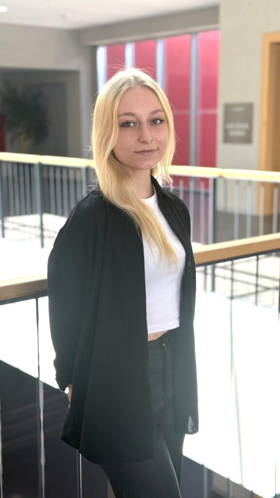 Eine Person mit langen blonden Haaren, die eine schwarze Jacke und ein weißes bauchfreies Top trägt, steht drinnen in der Nähe eines Geländers und lächelt sanft in die Kamera. Helles Licht durchflutet den Hintergrund und unterstreicht ihre selbstbewusste Ausstrahlung, während sie sich auf einen weiteren Tag ihres Bioanalytik-Studiums vorbereitet. at Hochschule Coburg
