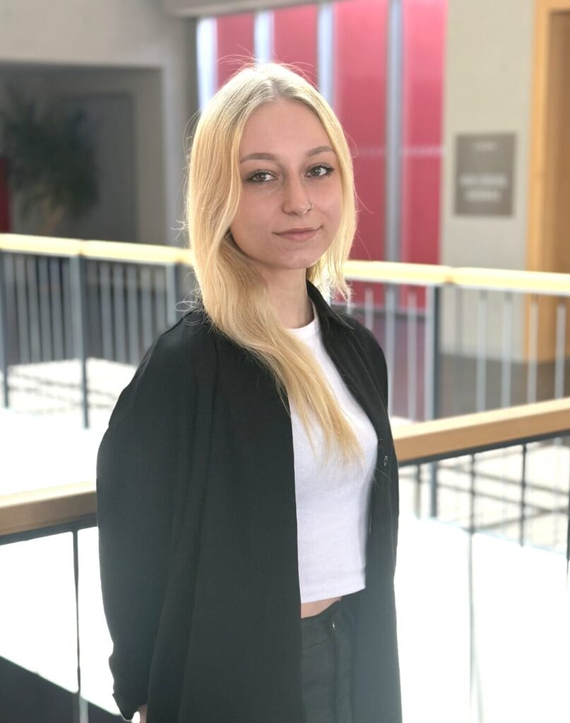 Eine Person mit langen blonden Haaren, die eine schwarze Jacke und ein weißes bauchfreies Top trägt, steht drinnen in der Nähe eines Geländers und lächelt sanft in die Kamera. Helles Licht durchflutet den Hintergrund und unterstreicht ihre selbstbewusste Ausstrahlung, während sie sich auf einen weiteren Tag ihres Bioanalytik-Studiums vorbereitet. at Hochschule Coburg