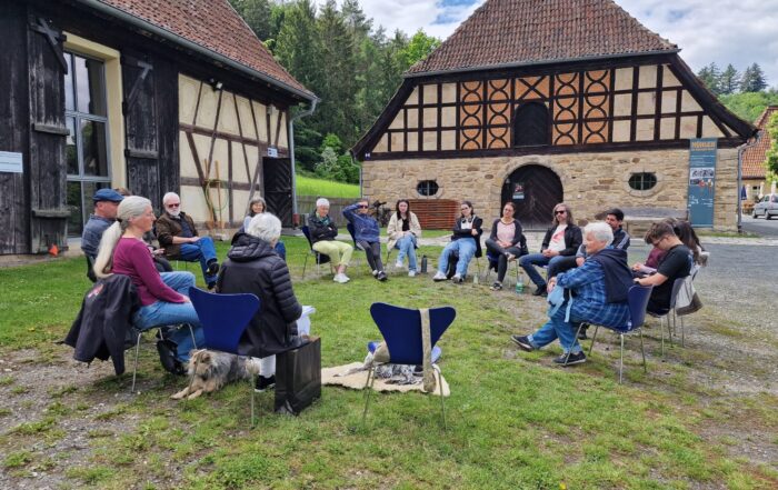 Eine Gruppe von Menschen sitzt im Freien im Kreis auf Stühlen, unterhält sich und hört zu. Sie befinden sich zwischen zwei rustikalen Gebäuden mit Fassaden aus Holz und Stein. Um sie herum erstrecken sich grünes Gras und Bäume. at Hochschule Coburg